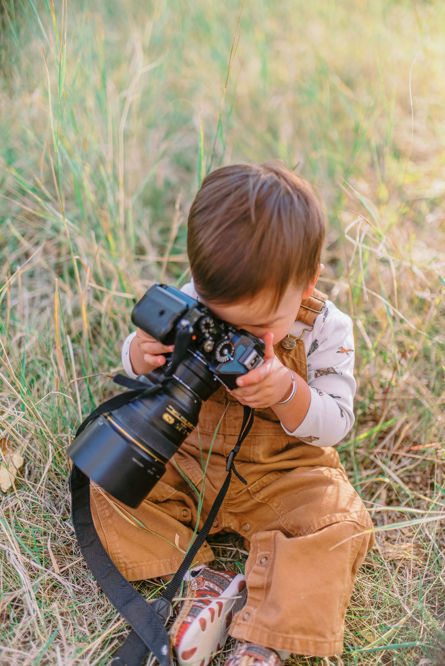 A young photographer in a brown jacket examines a professional camera while sitting in tall grass during golden hour.