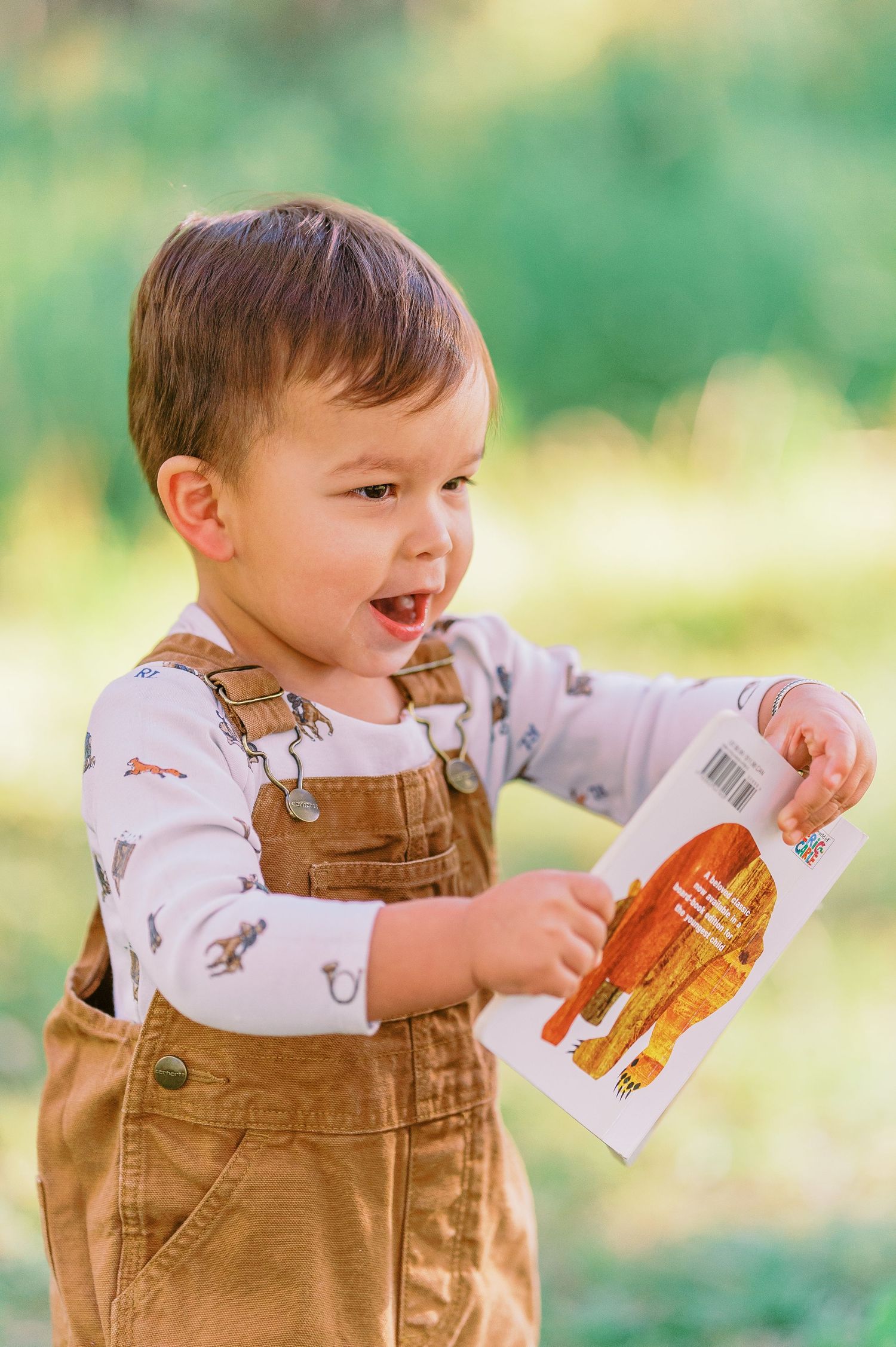 A small child in brown overalls plays with a book in a sunny outdoor setting.