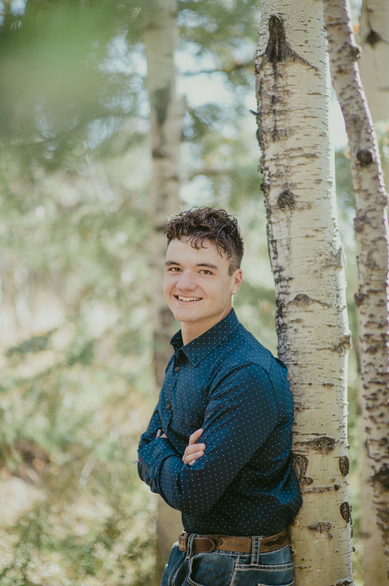 Young man in navy blue shirt smiling with arms crossed, leaning against birch trees in a forest setting.
