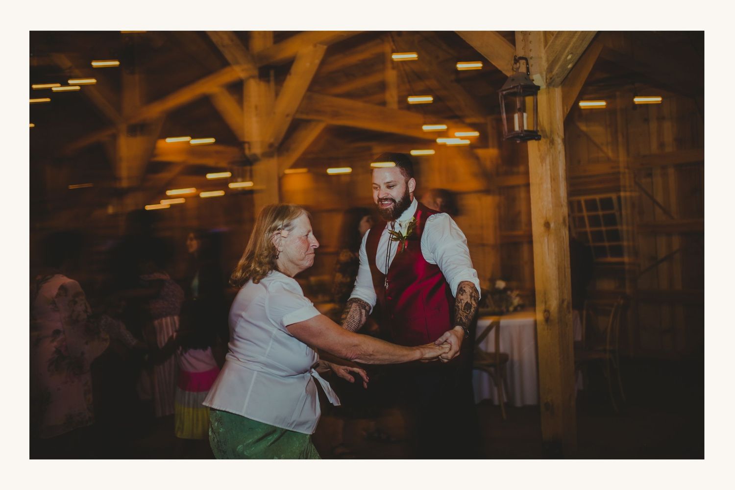 Groom in burgundy vest dancing with elderly woman at rustic barn wedding reception with warm string lights.