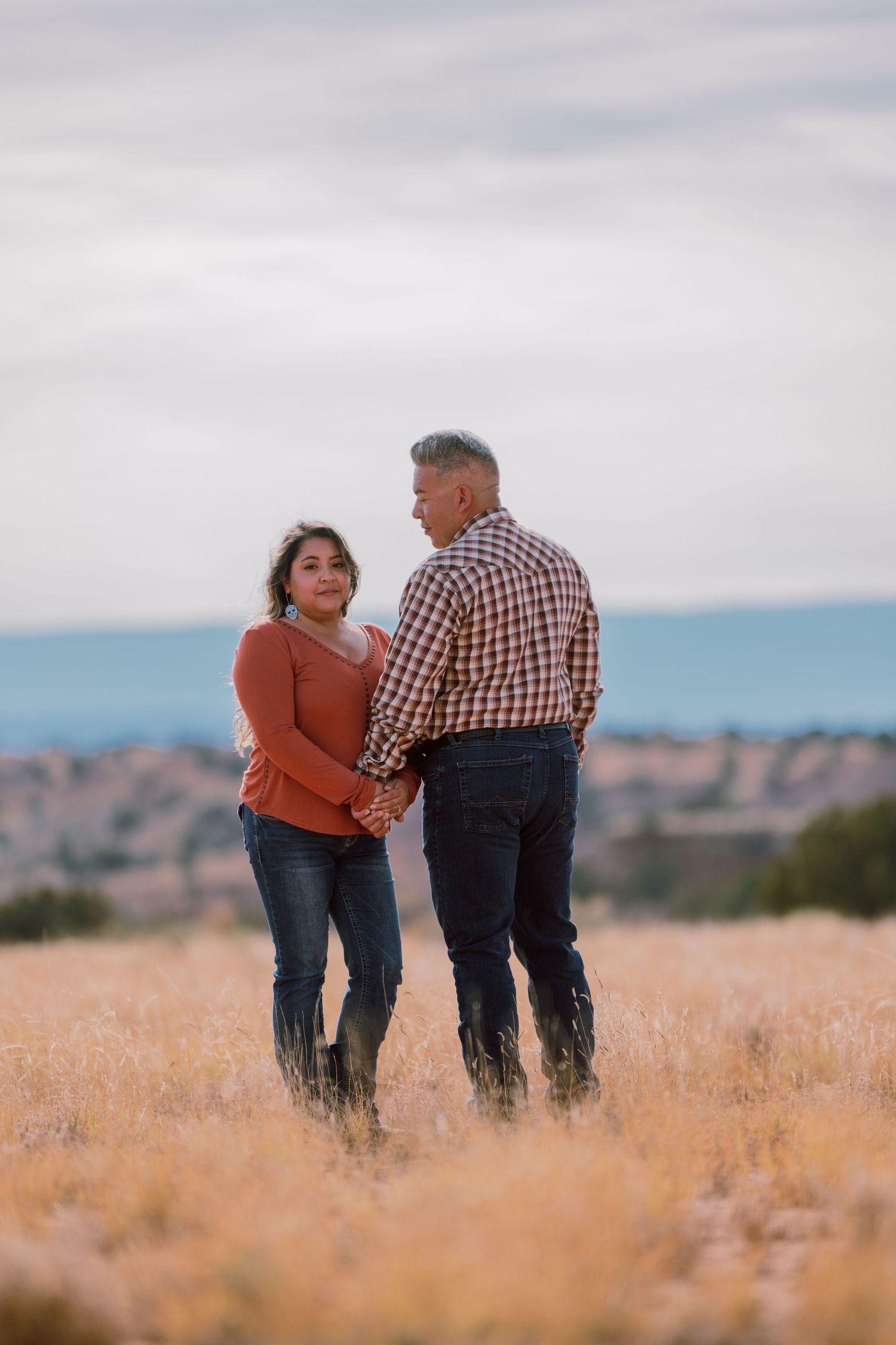 Couple holding hands and walking together through a golden field with hazy mountains in the background.