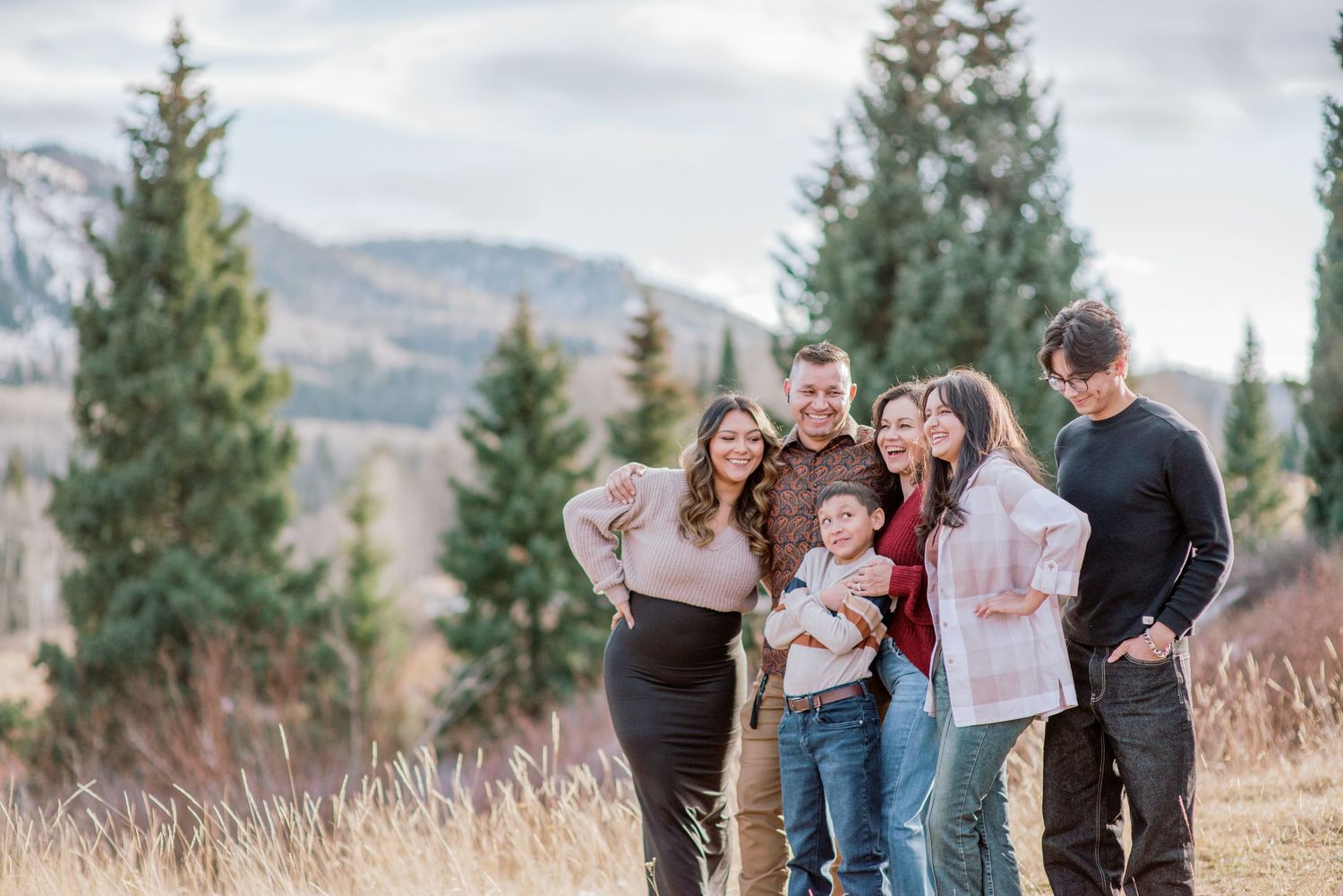 A group shares joyful laughter together outdoors among pine trees and mountains on an overcast day.