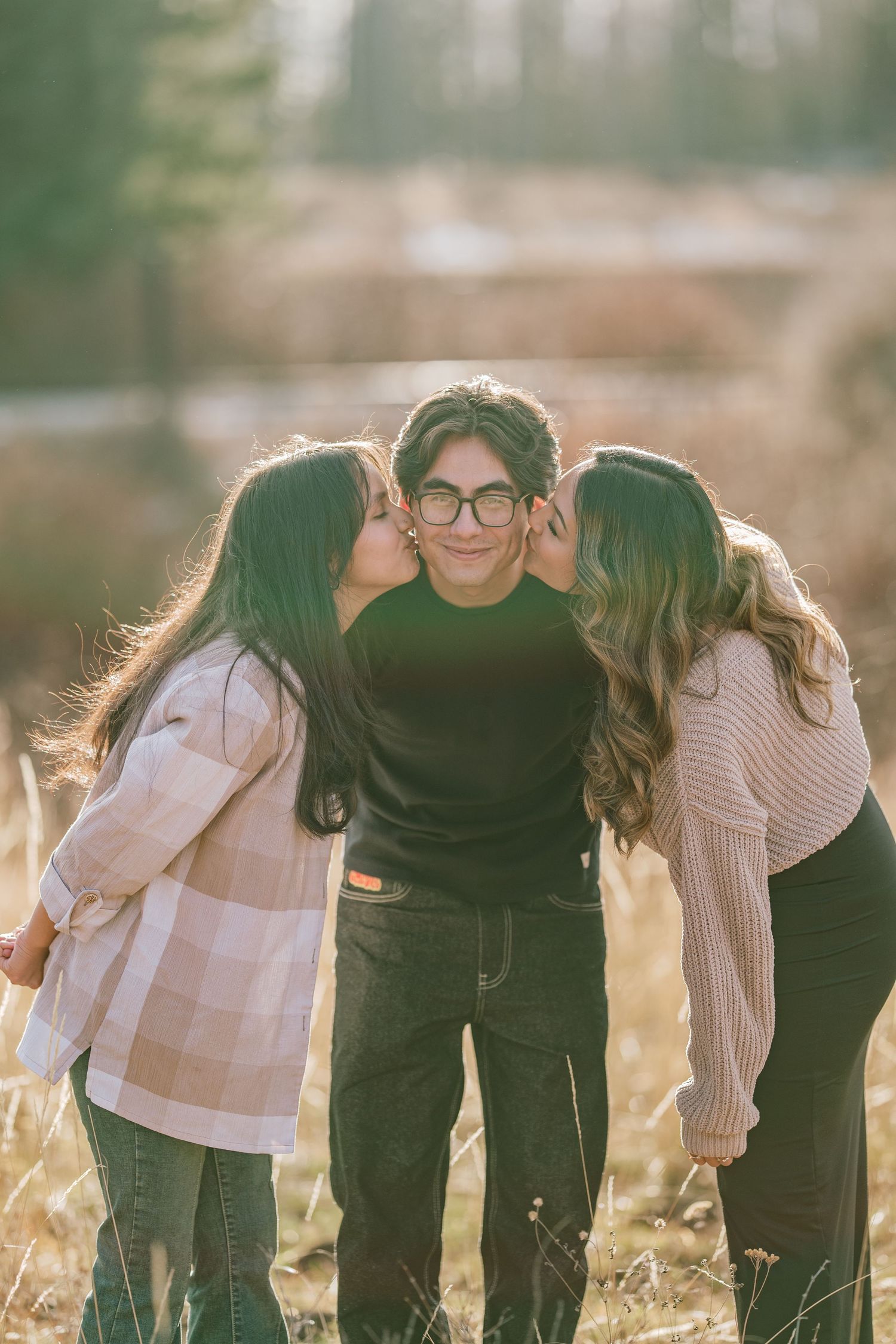 Three friends share a tender moment outdoors in golden sunlight as two lean in to kiss their companion's cheeks.