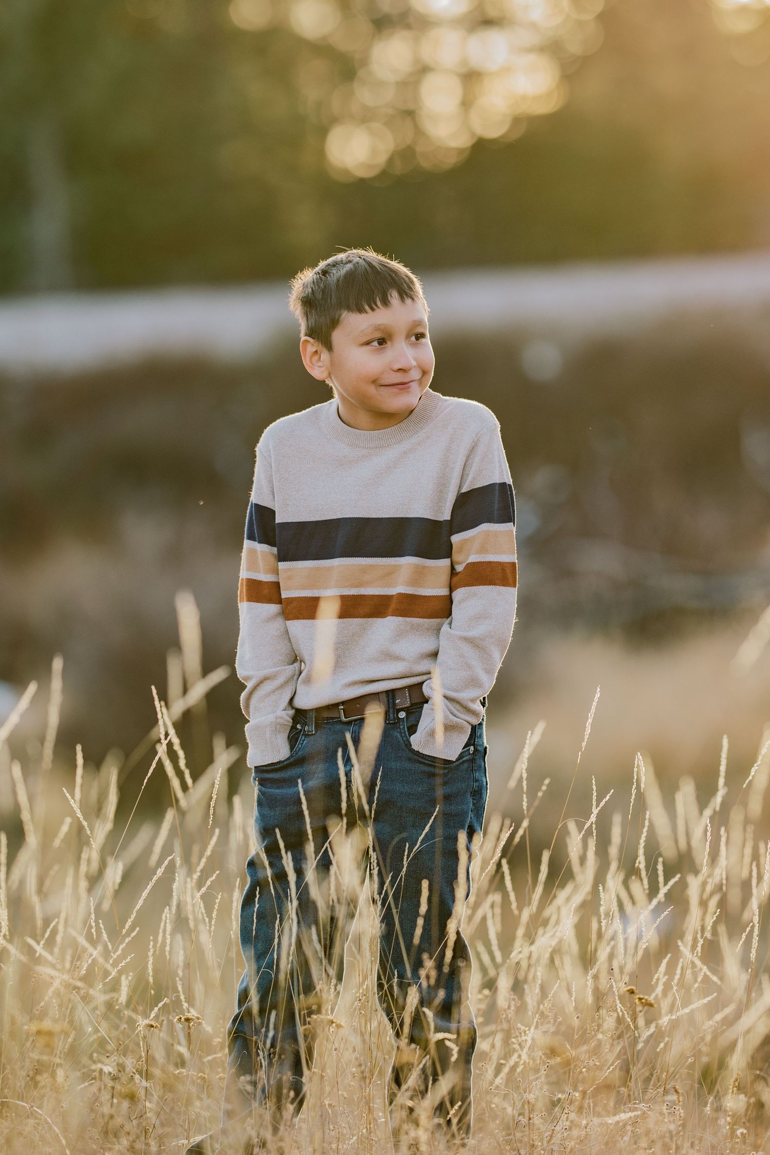A figure stands in a golden wheat field at sunset wearing a striped sweater and jeans, with soft bokeh lighting in background.