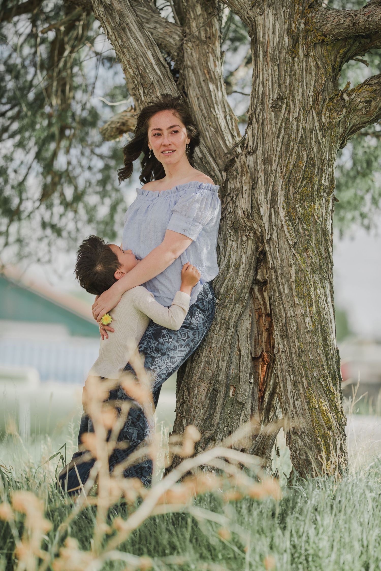 A tender moment shared between parent and child beneath a large tree in a dreamy outdoor setting with tall grass.