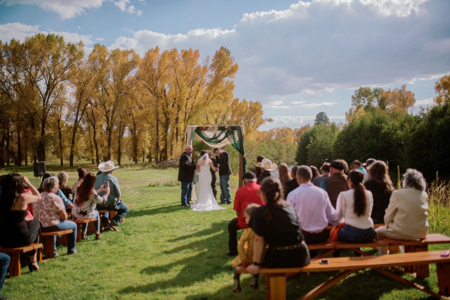 Outdoor wedding ceremony with wooden bench seating and golden autumn trees in background.