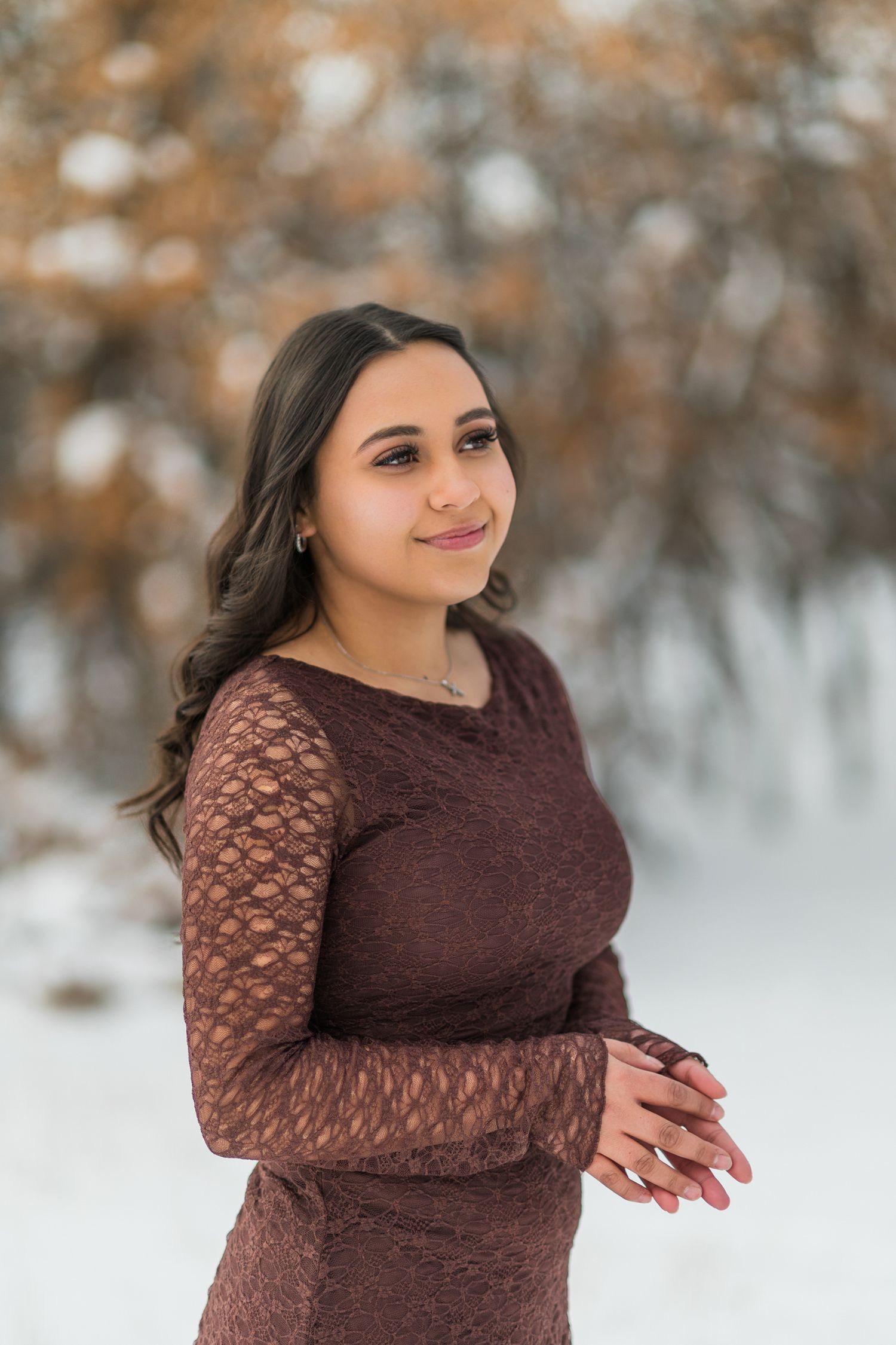 A woman in a brown lace dress poses outdoors in winter with snow and autumn trees in the background.