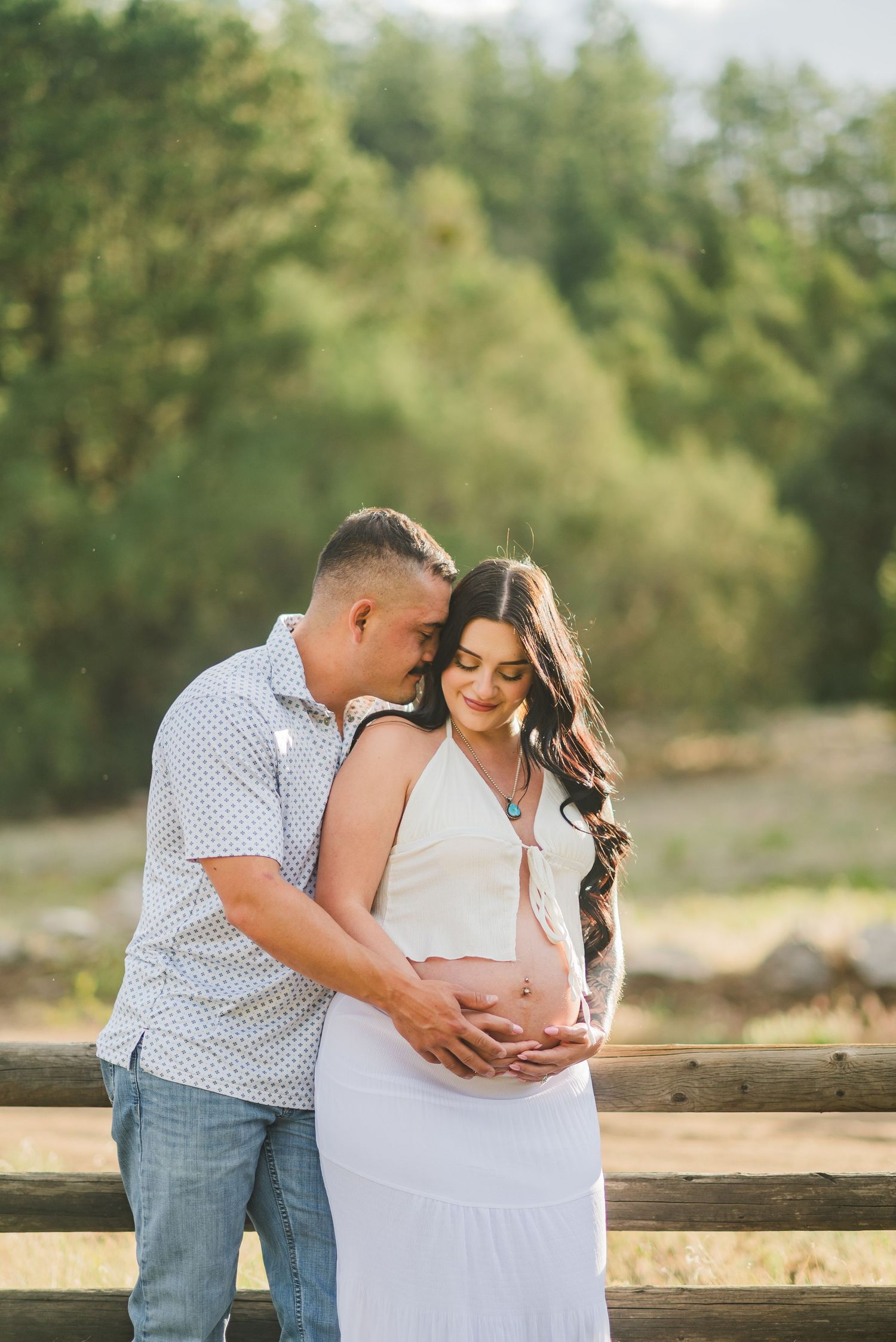 Couple tenderly cradling pregnant belly outdoors near rustic fence, surrounded by lush green trees in warm sunlight.