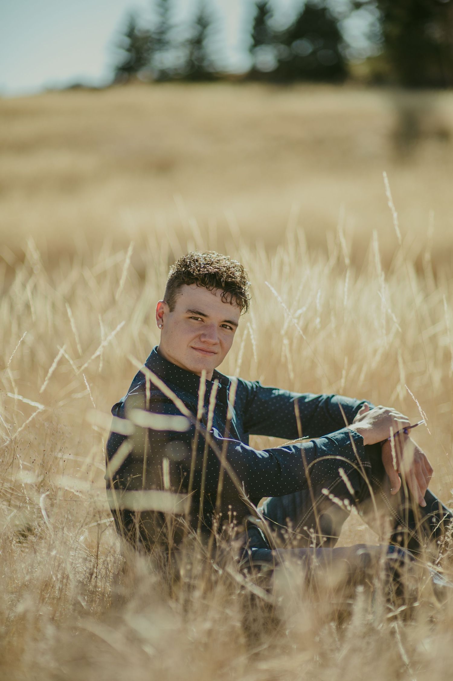 Young man sitting in golden dry grass field, smiling, wearing dark sweater, with trees on horizon on sunny day.