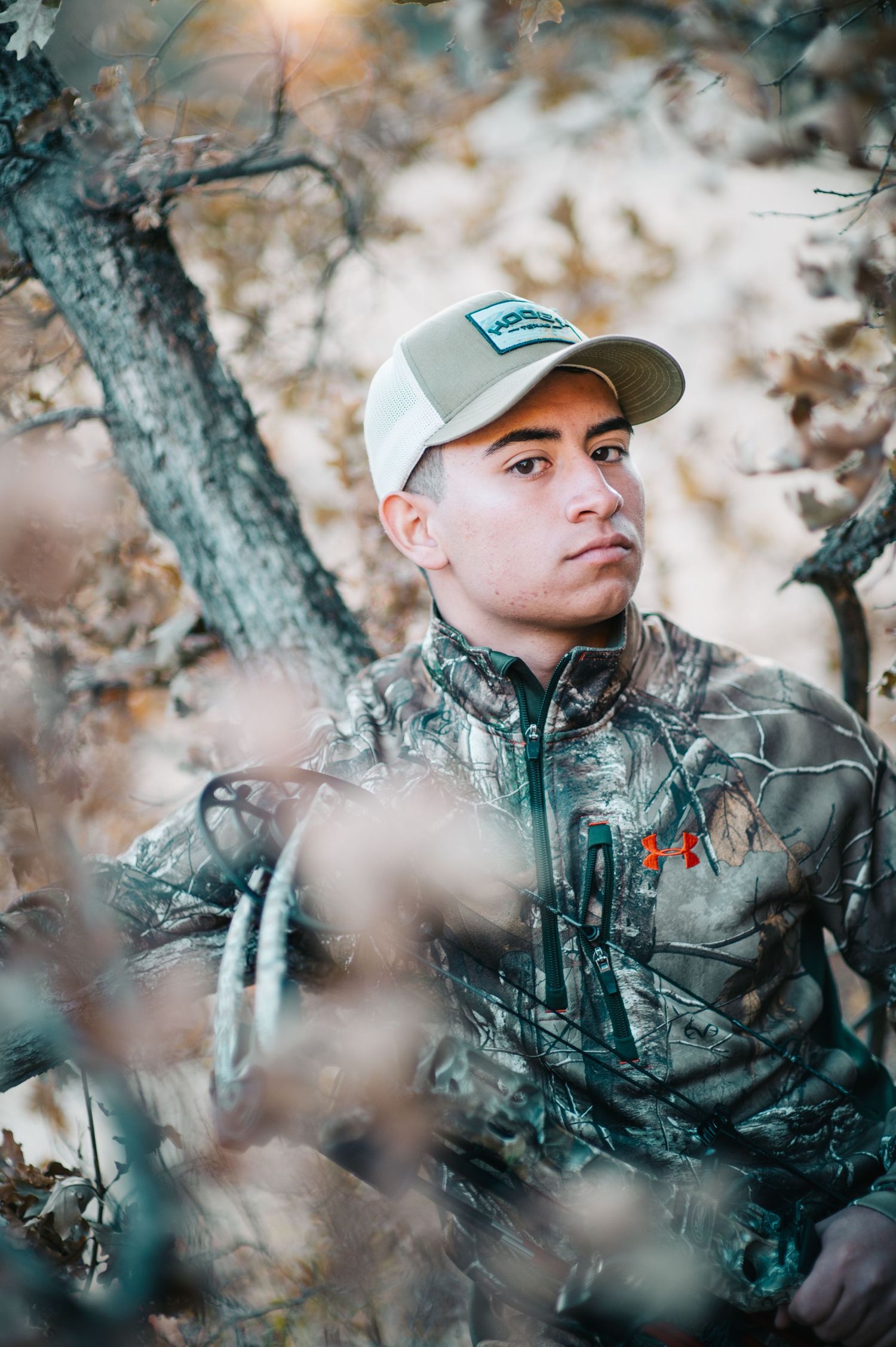 Young hunter in camouflage jacket and cap standing among bare autumn trees outdoors.