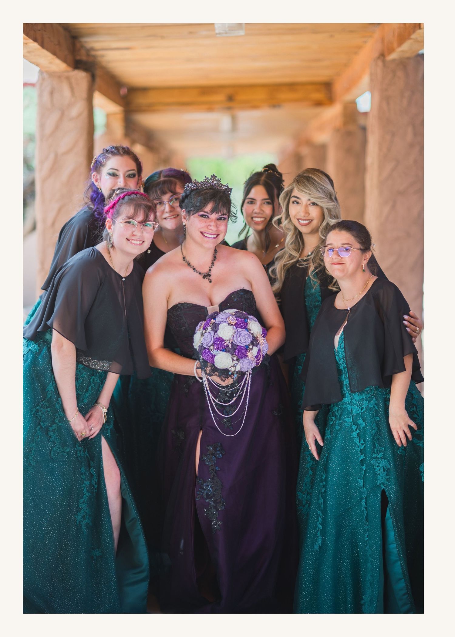Bride in purple gown holds bouquet surrounded by bridesmaids in teal and black dresses at an outdoor wedding.