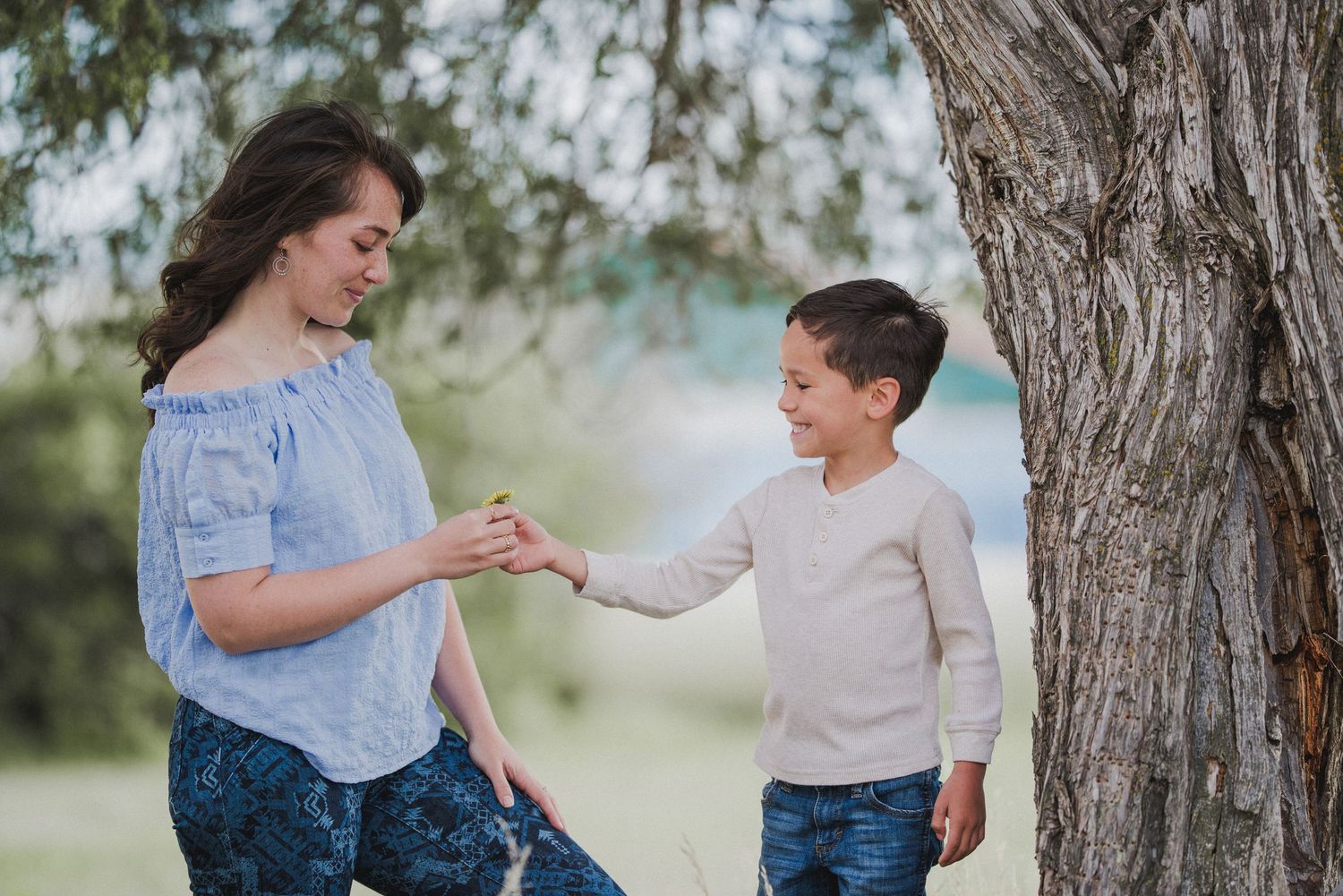 Two people communicate with hand gestures while standing next to a tree trunk in a natural outdoor setting.