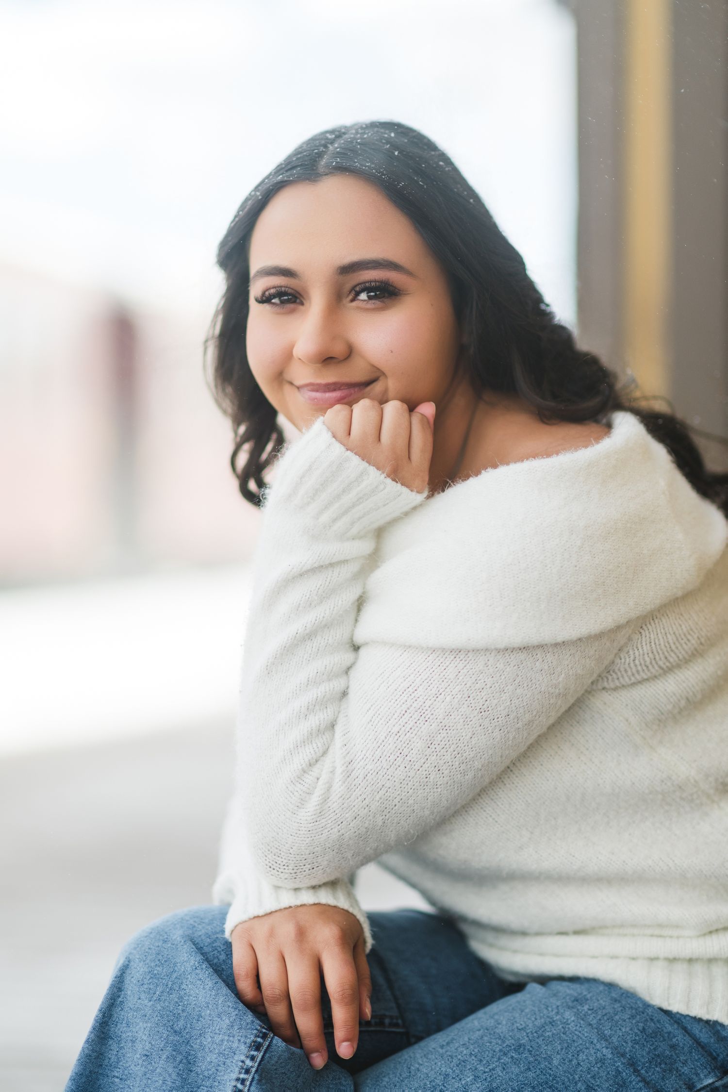 Woman in cozy white turtleneck sweater and jeans smiles warmly while sitting in natural lighting.