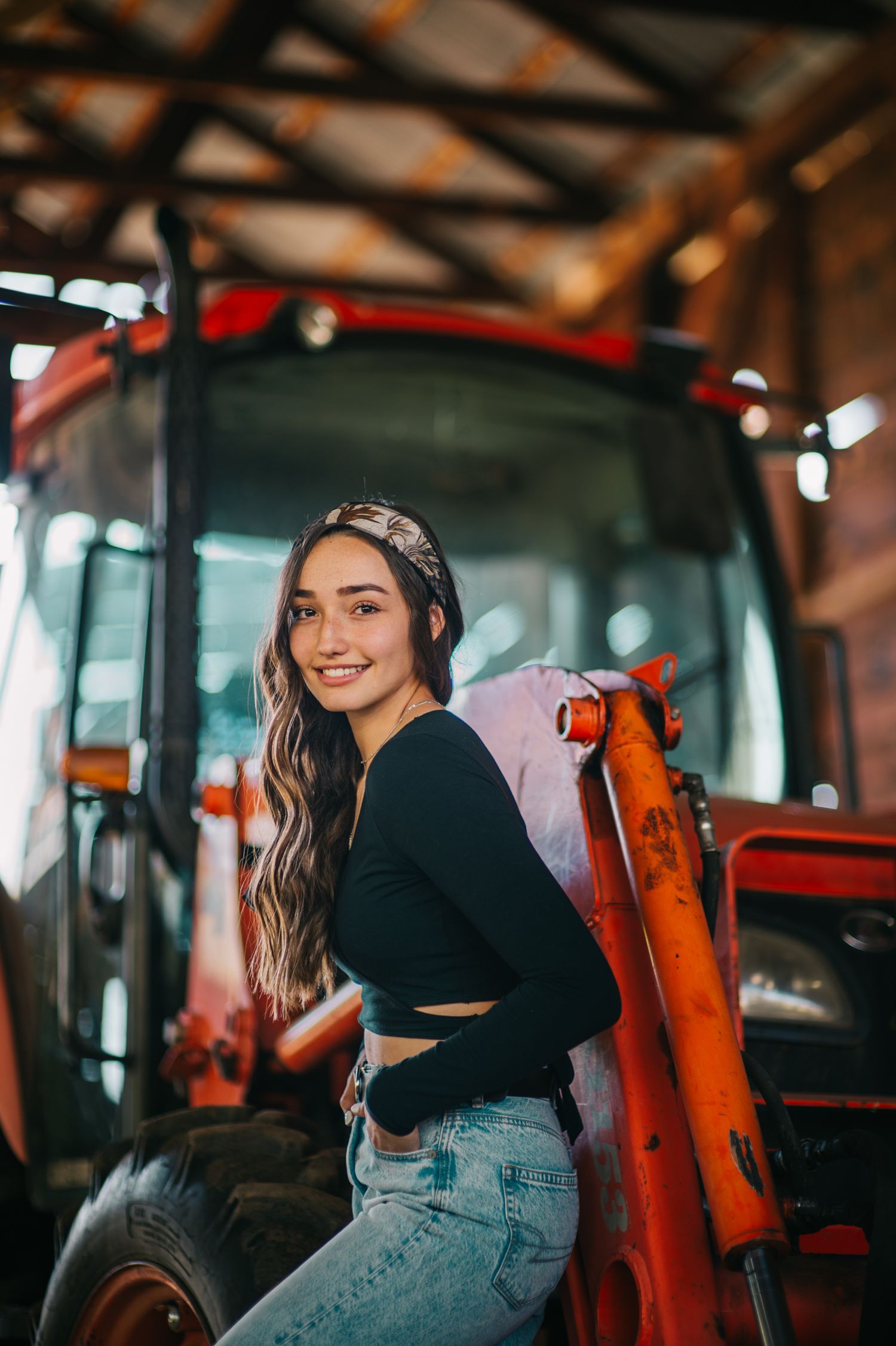 Model poses on orange tractor in black crop top and blue jeans inside barn setting.