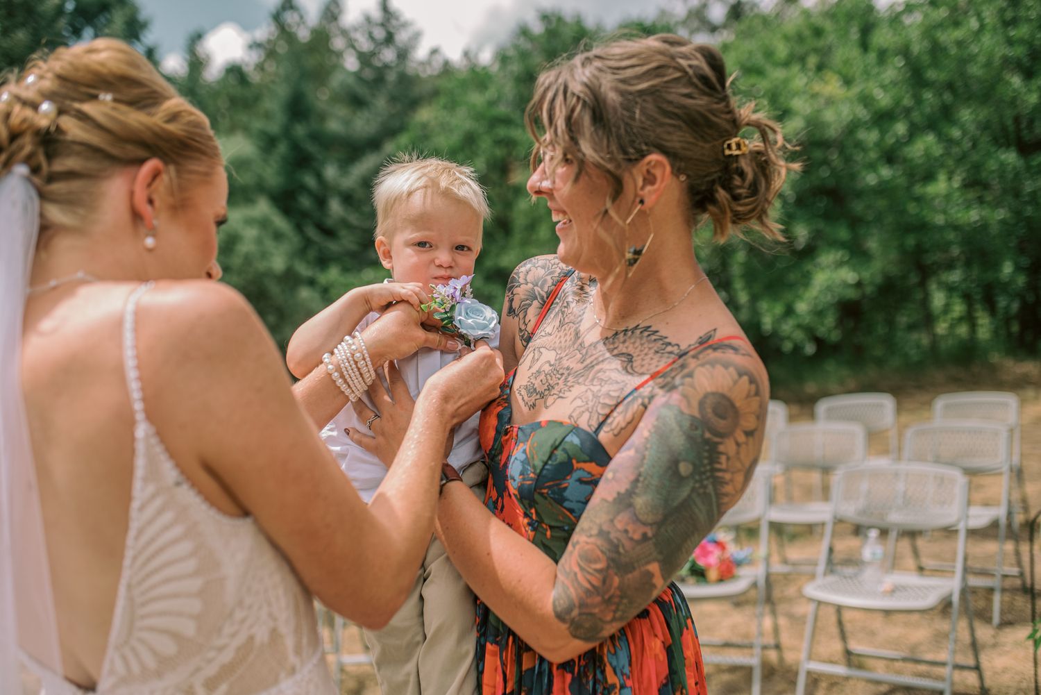 Two women in wedding attire hold a baby outdoors at a ceremony, one with colorful tattoos, surrounded by white chairs.
