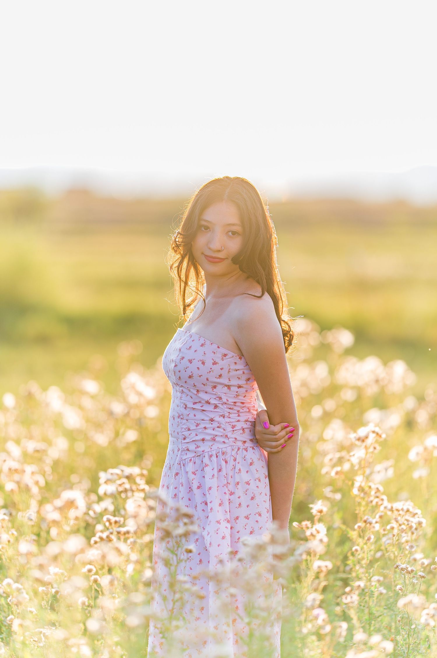 A person in a floral dress poses in a sunlit wildflower field during golden hour.