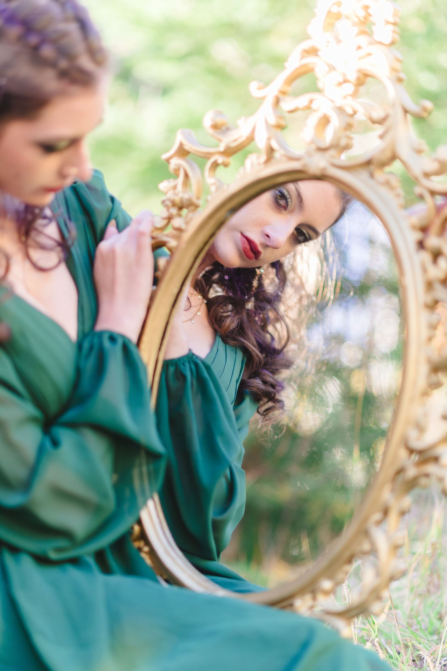 A person in an emerald green dress poses with an ornate gold mirror in a dreamy outdoor setting.