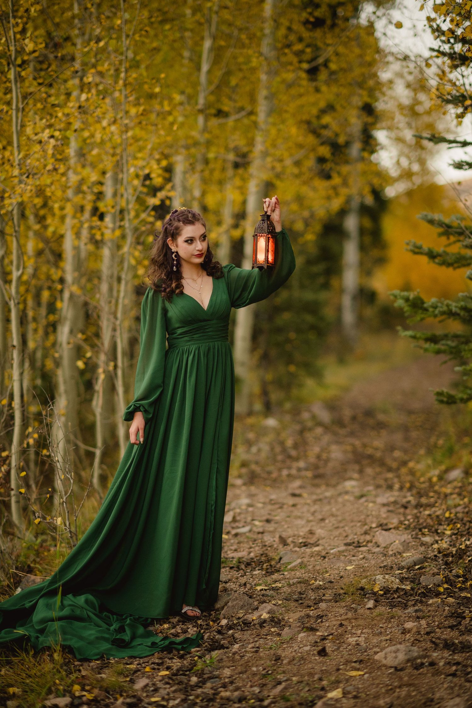 Woman in flowing emerald green dress poses gracefully among golden autumn aspen trees on forest path.