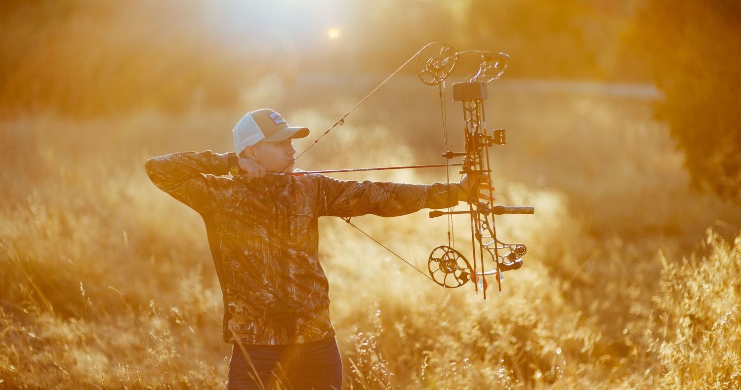 Archer in camouflage aiming compound bow in golden sunlit field during bowhunting season.