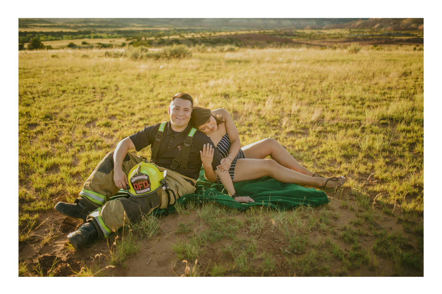 Couple relaxing in a sunny field, man in firefighter gear, woman in striped outfit, sitting on green blanket.