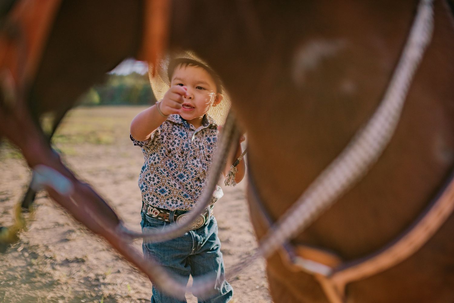 Young child in cowboy hat smiling between two brown horses on a ranch, holding reins outdoors.