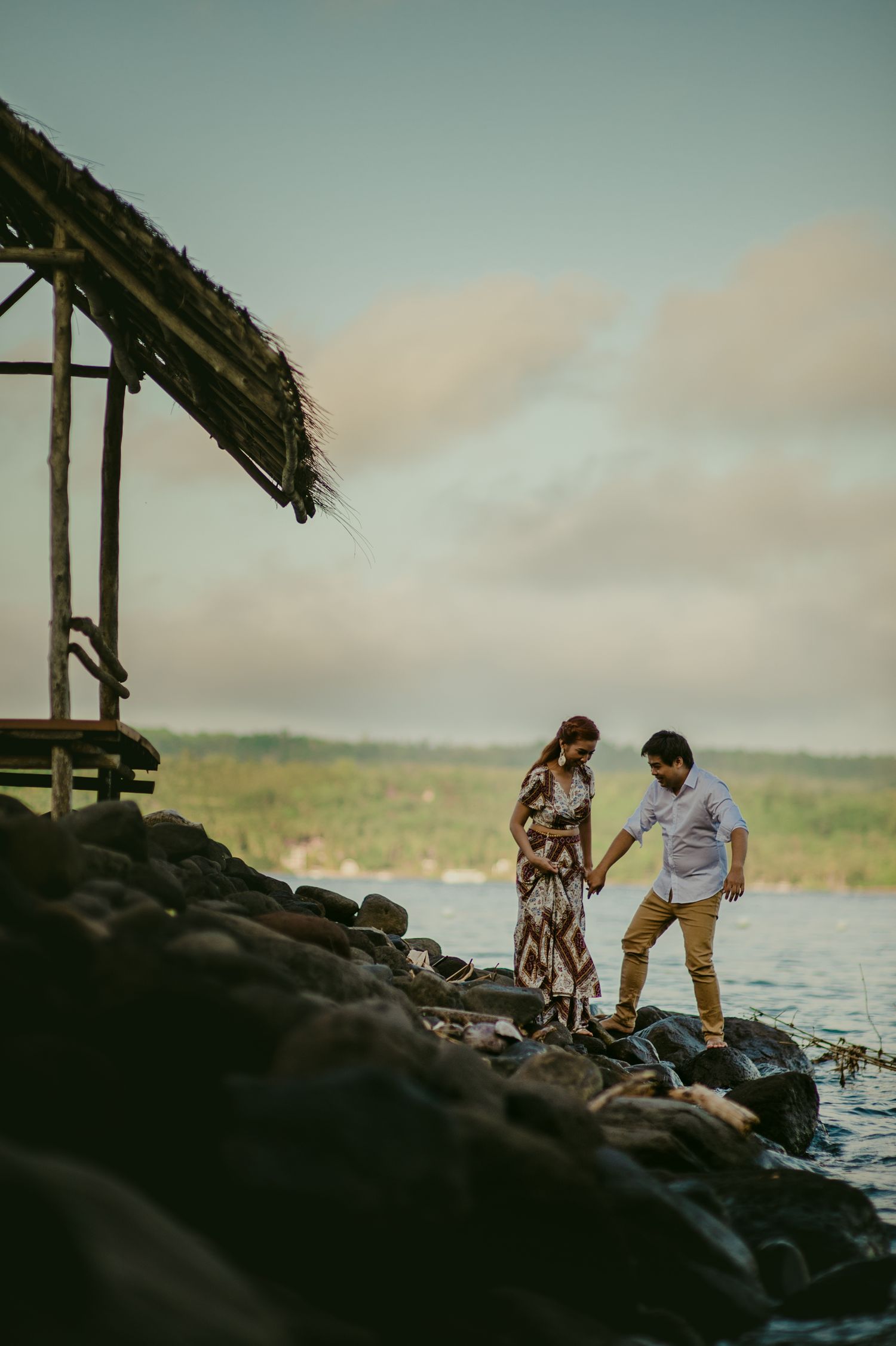 A couple exploring rocky shoreline beneath a rustic wooden structure near the ocean.