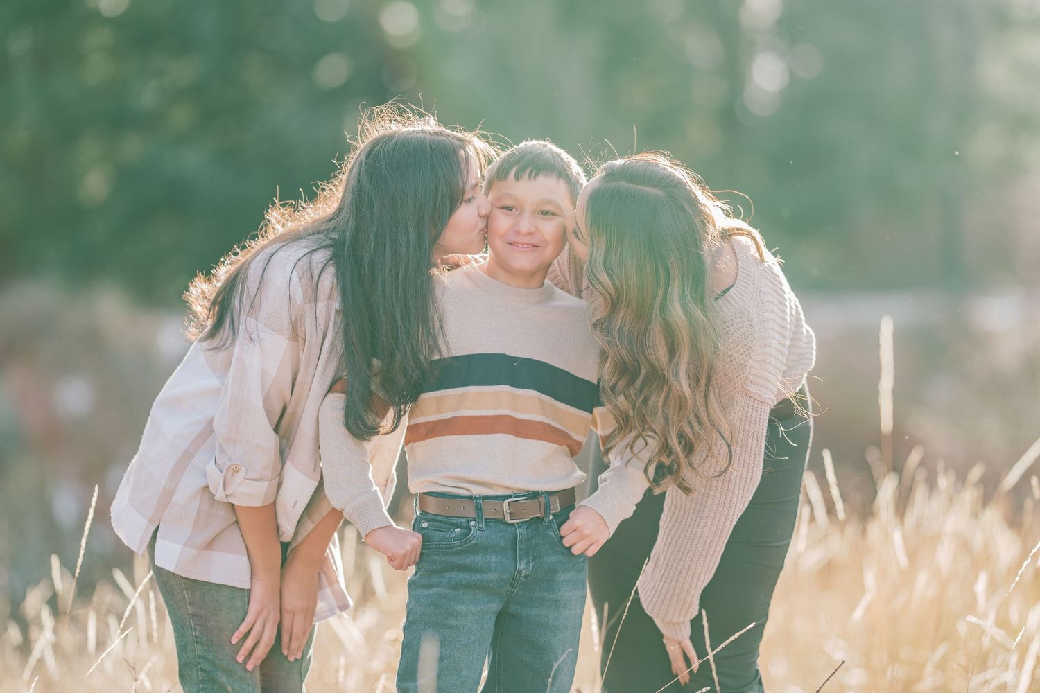 Three friends share an affectionate moment together in a sun-lit field with golden wheat grass and green trees.