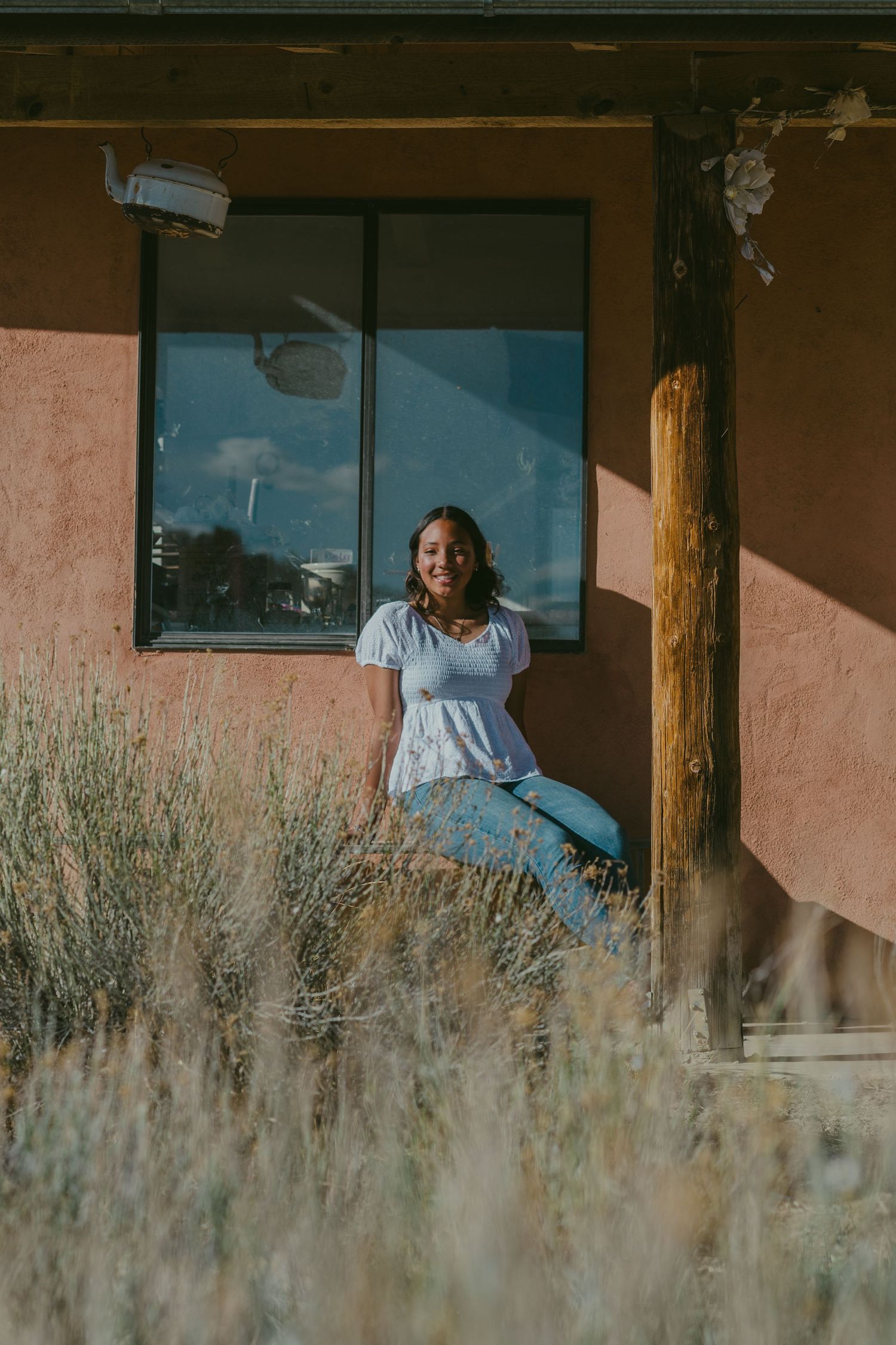 A person in a white top and jeans sits by a window of a rustic adobe building with dry grass in the foreground.