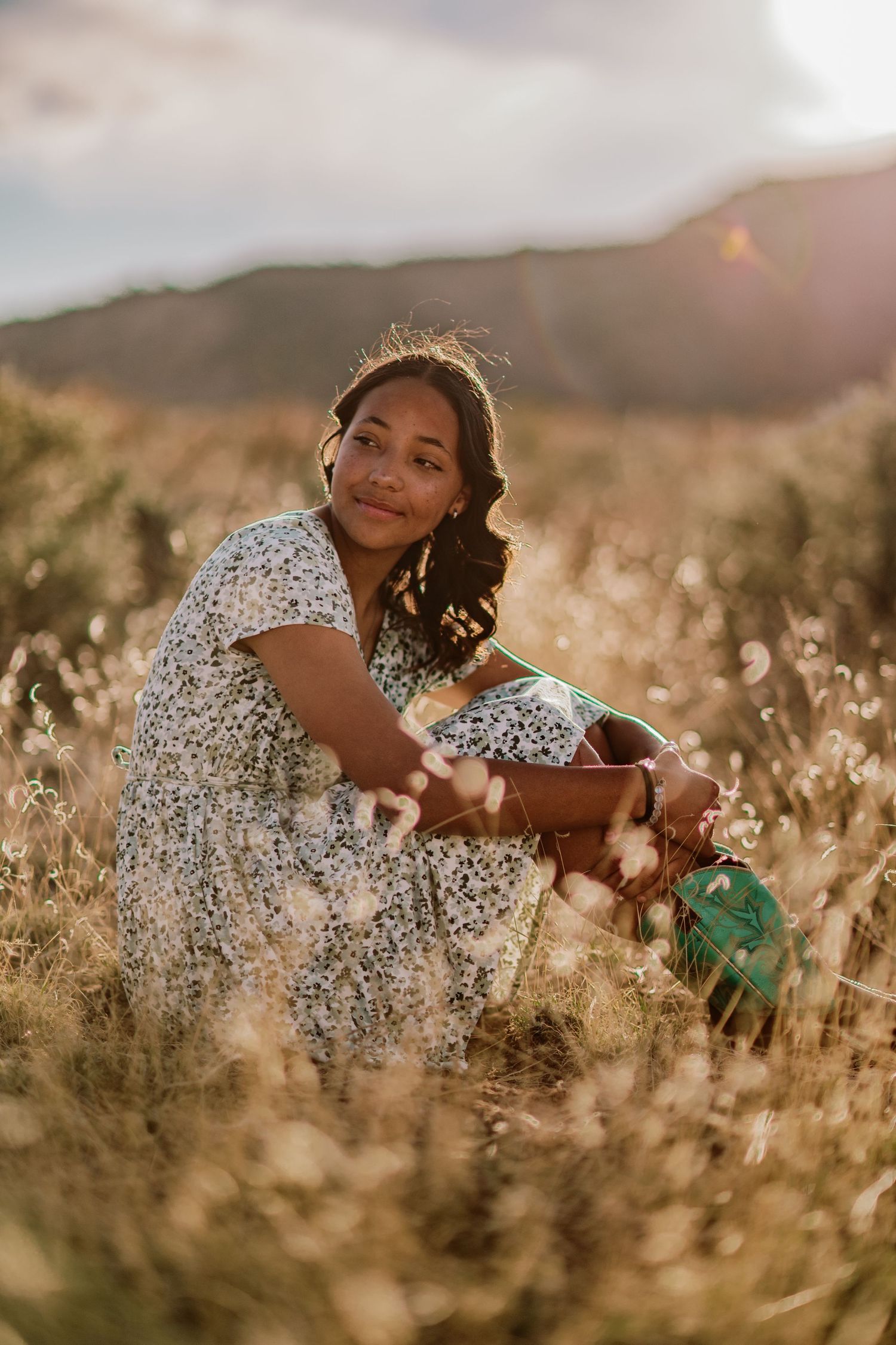 A person in a floral dress sits thoughtfully in a golden sunlit field of tall grass during sunset.