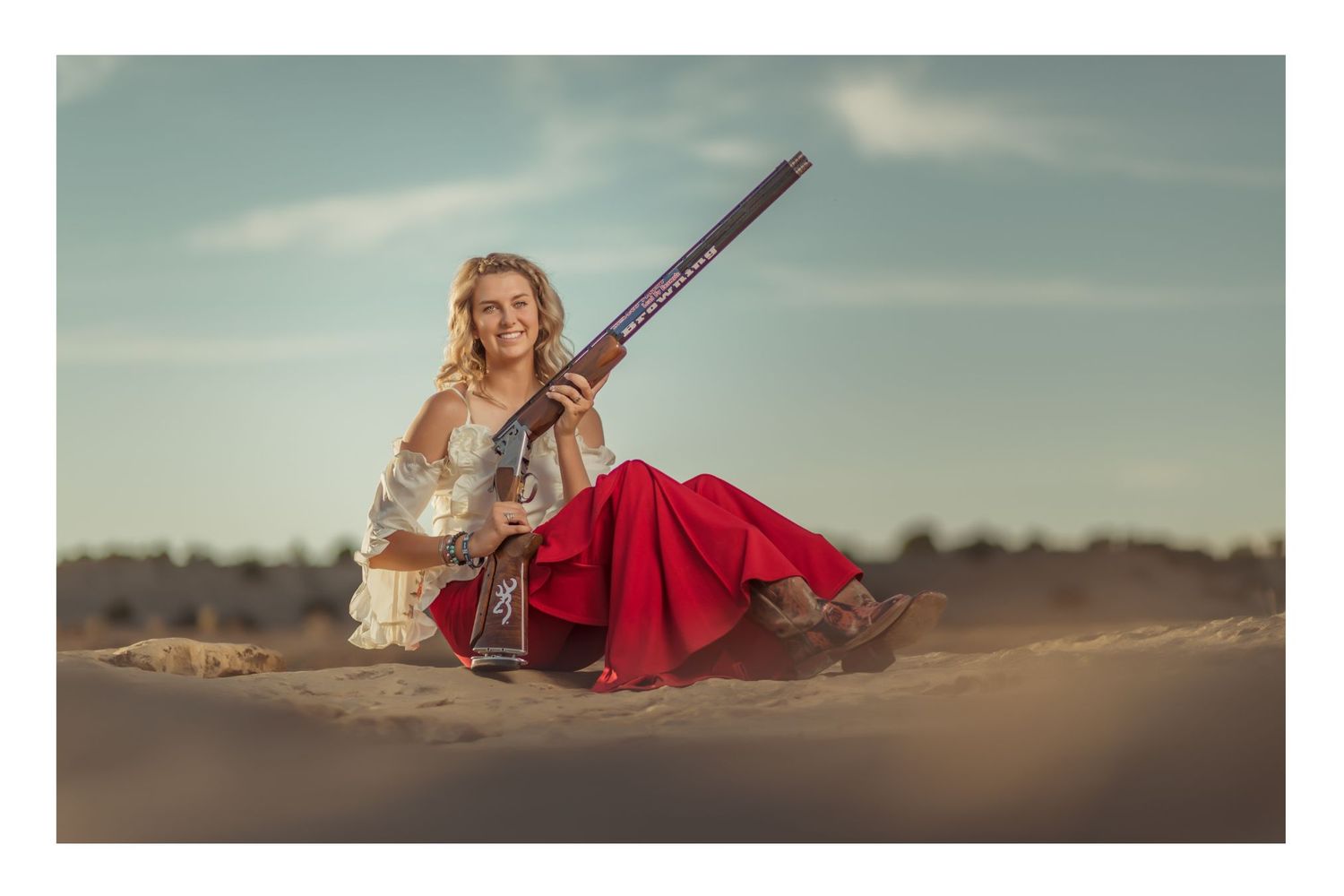 Woman in red skirt and white blouse poses with a musket rifle on sandy dunes under a blue sky.