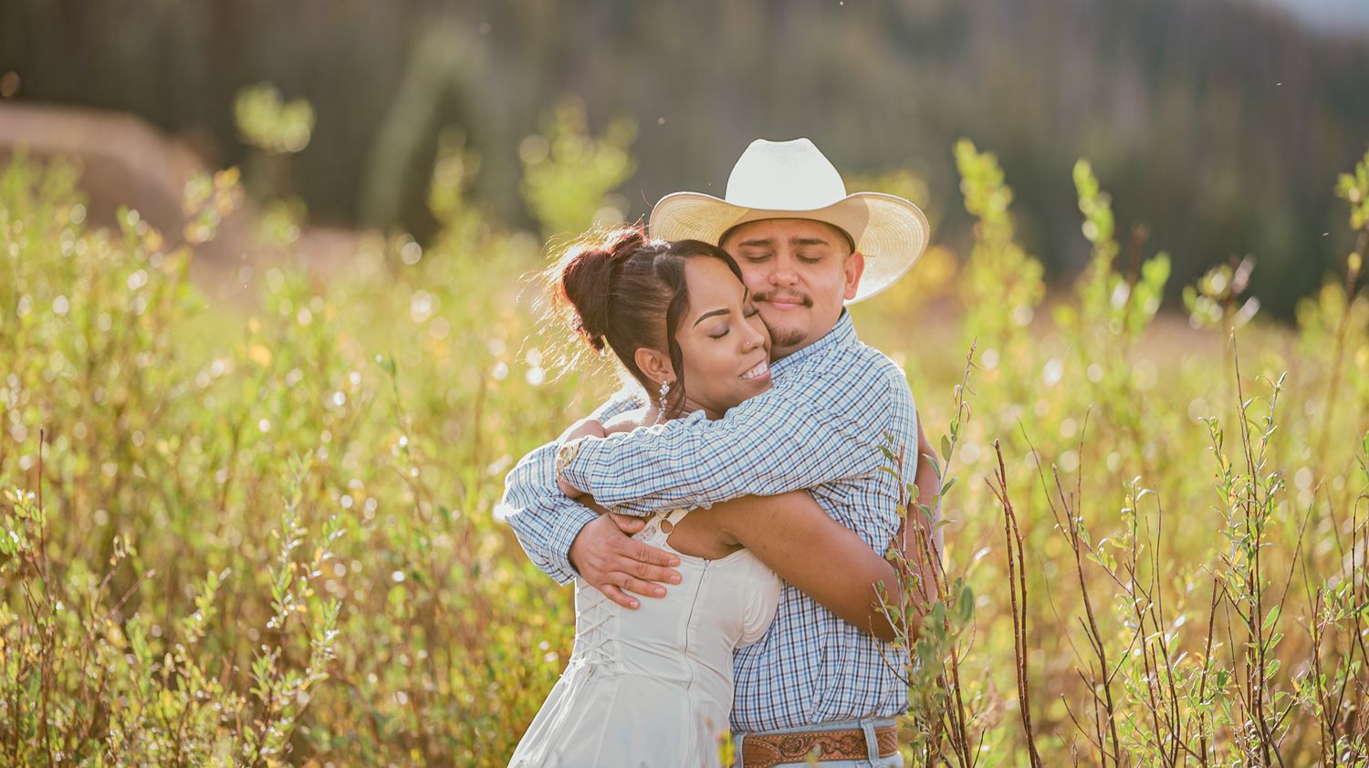 Couple embracing in a sunny meadow, man wearing a cowboy hat and blue shirt, woman in white dress.