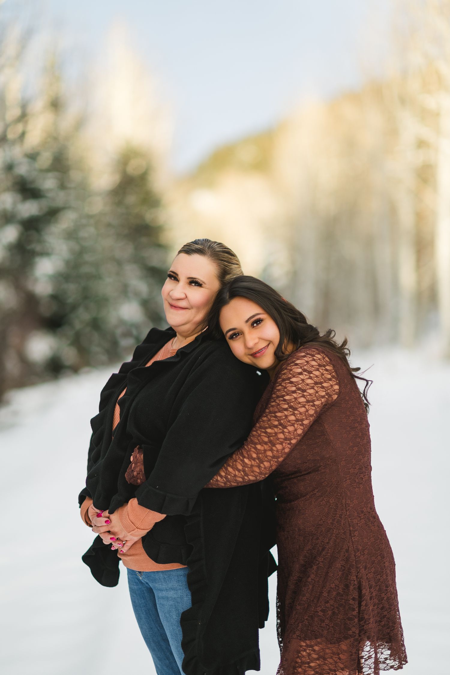 Two people embrace while standing in a snowy winter landscape with trees in the background.