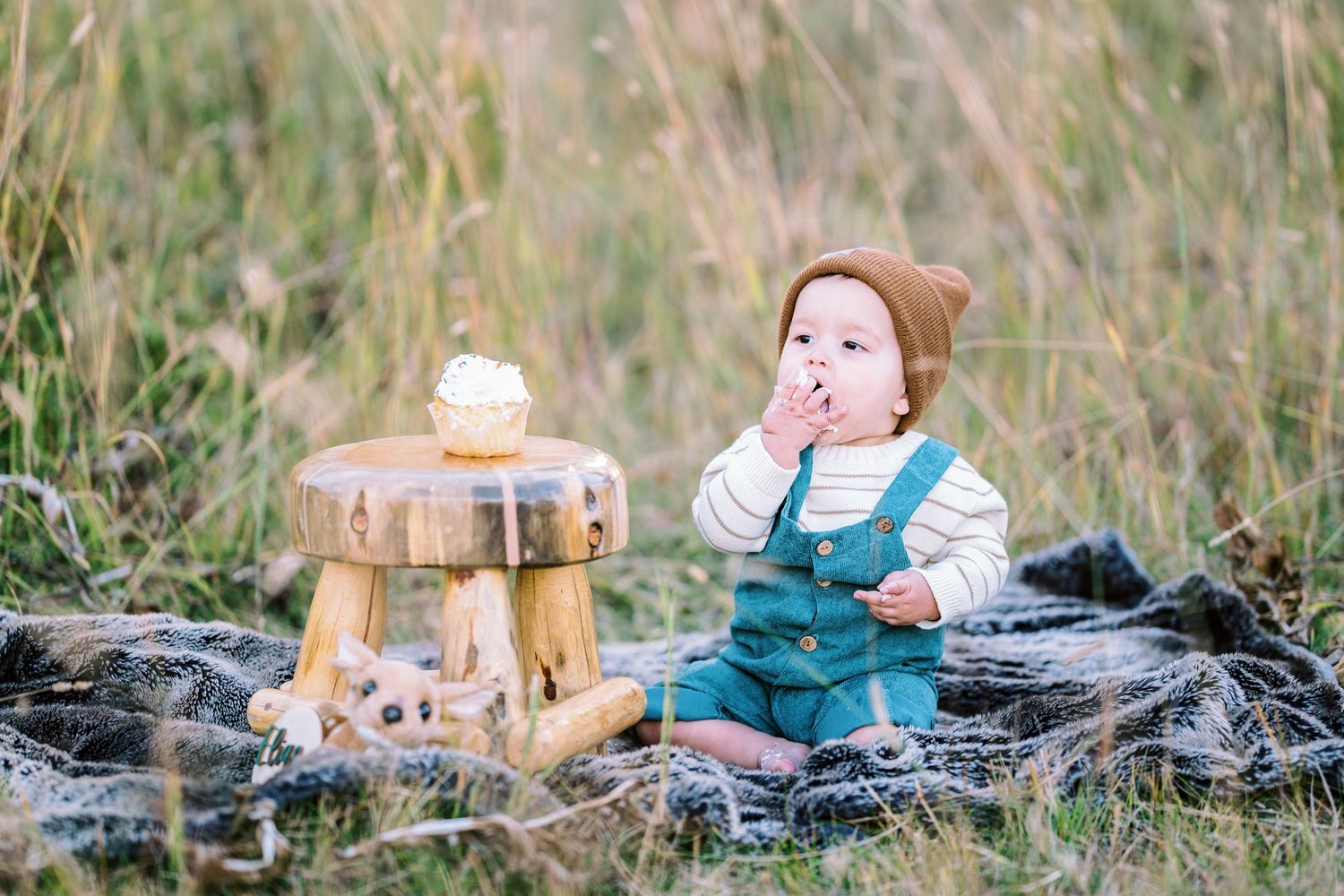 A baby in overalls enjoys snacks at a small wooden table in an outdoor setting.