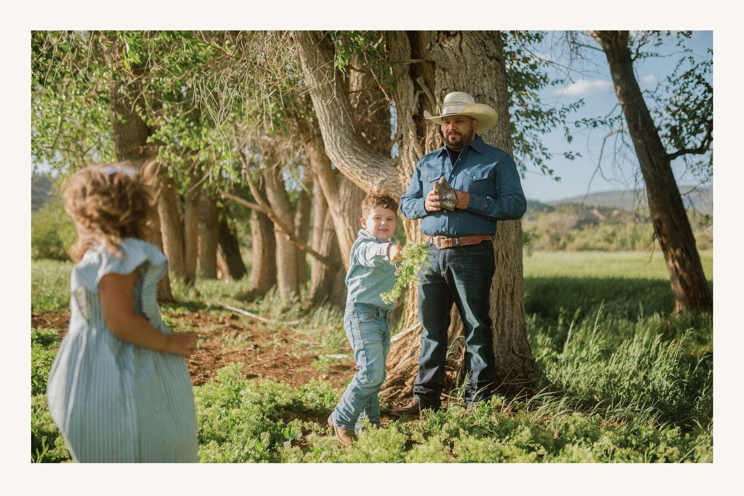 A man in a cowboy hat leans against a tree in a lush field, holding a drink while a child and woman in blue stand nearby.