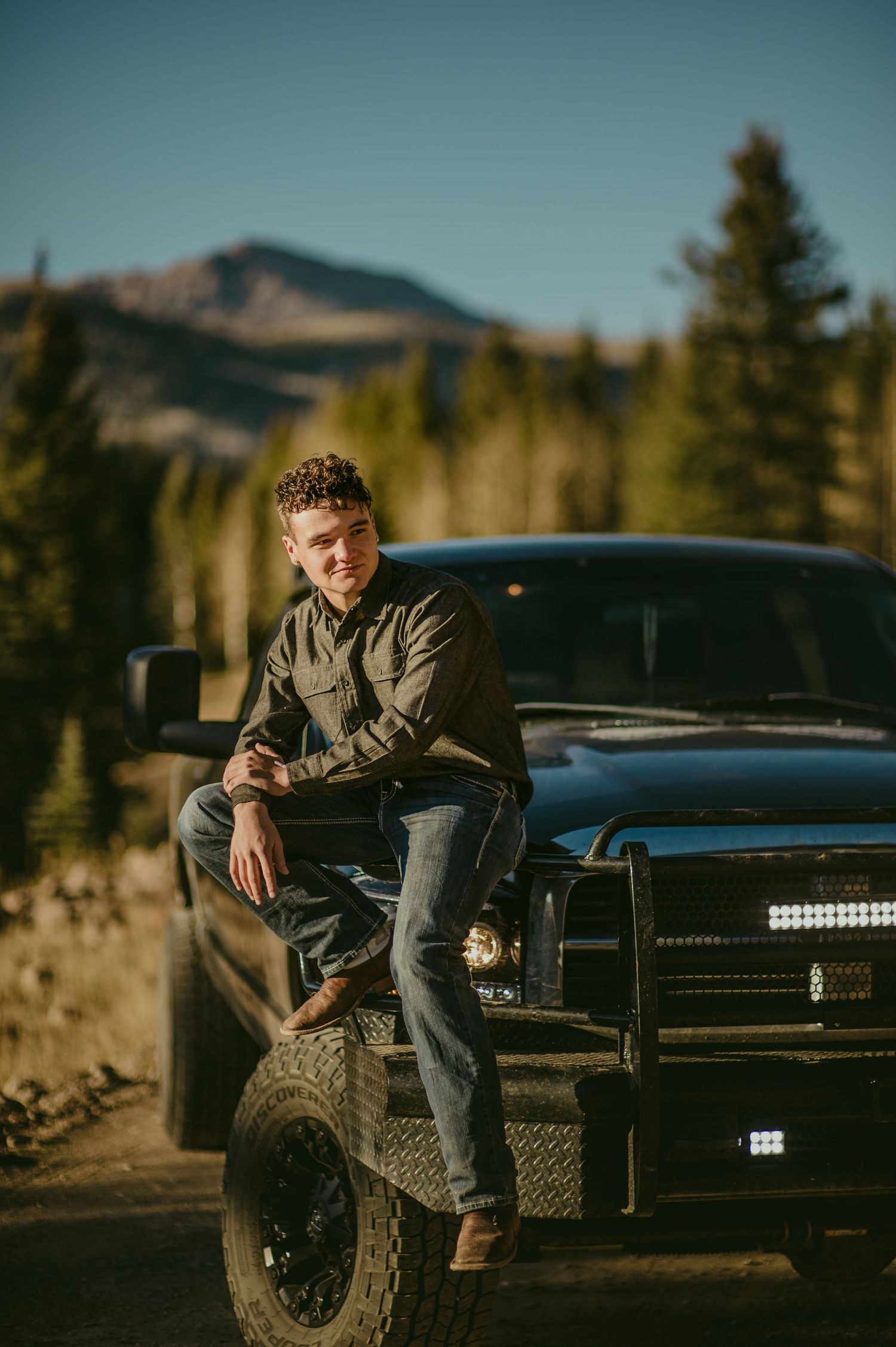 A young man leans confidently on a black truck bumper with a mountain forest backdrop.