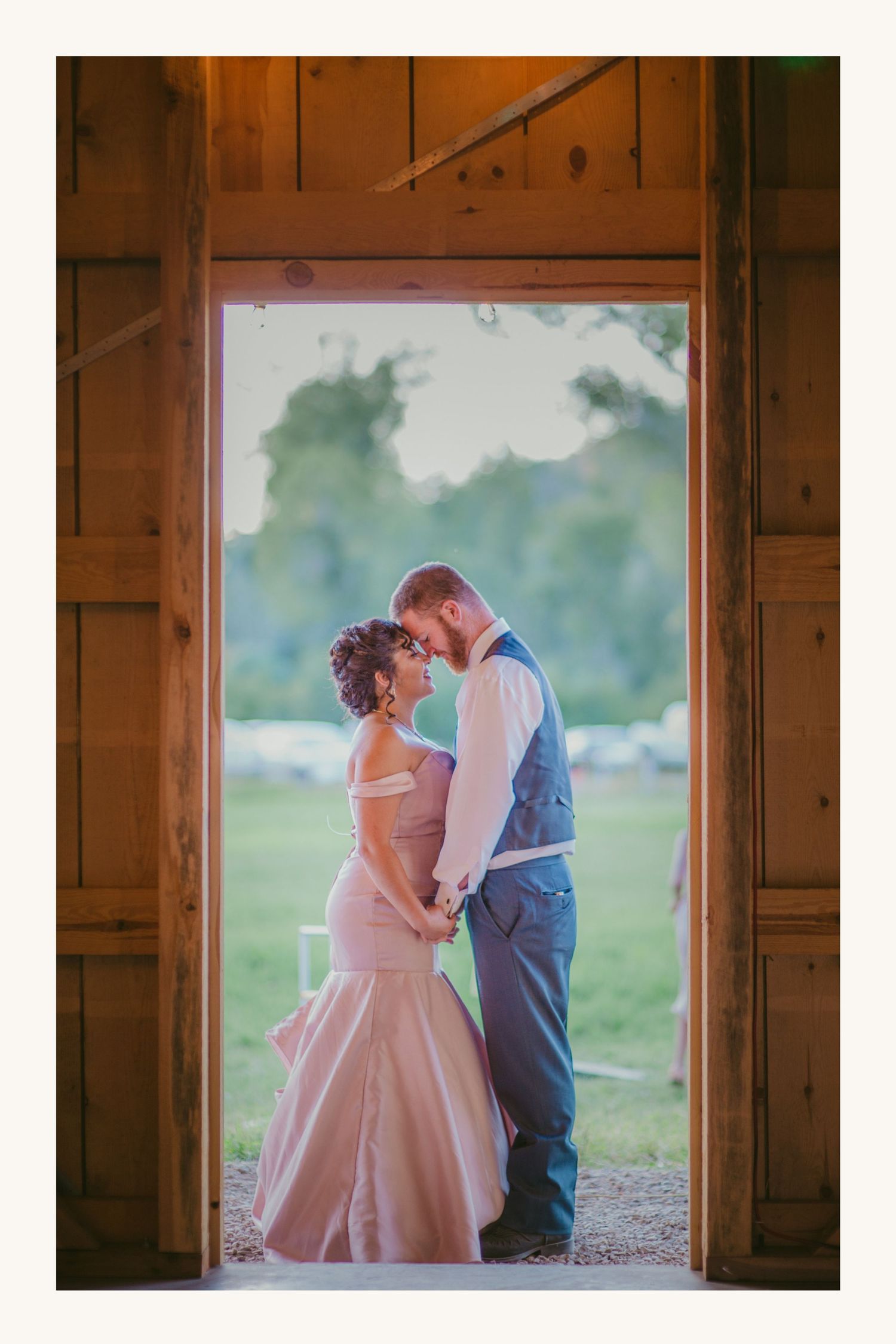 Couple sharing romantic moment in barn doorway, bride in pink mermaid gown, groom in blue vest, rustic outdoor wedding.