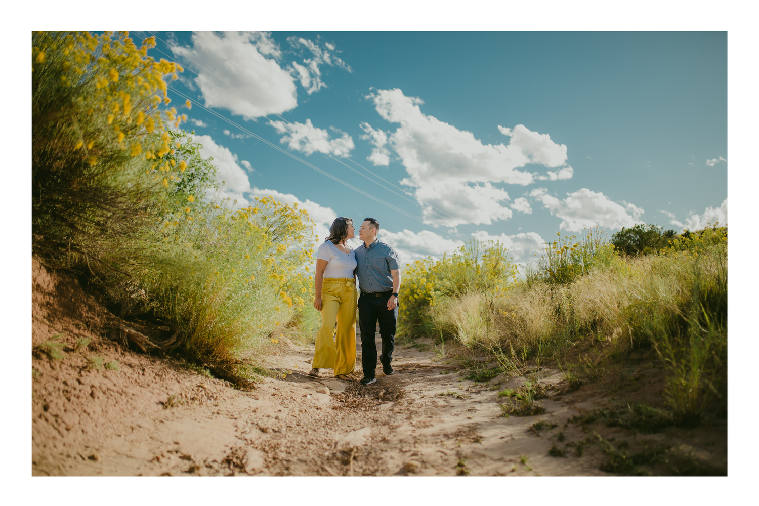 Couple walking on sandy desert path surrounded by green shrubs and yellow wildflowers under a bright blue sky with clouds.