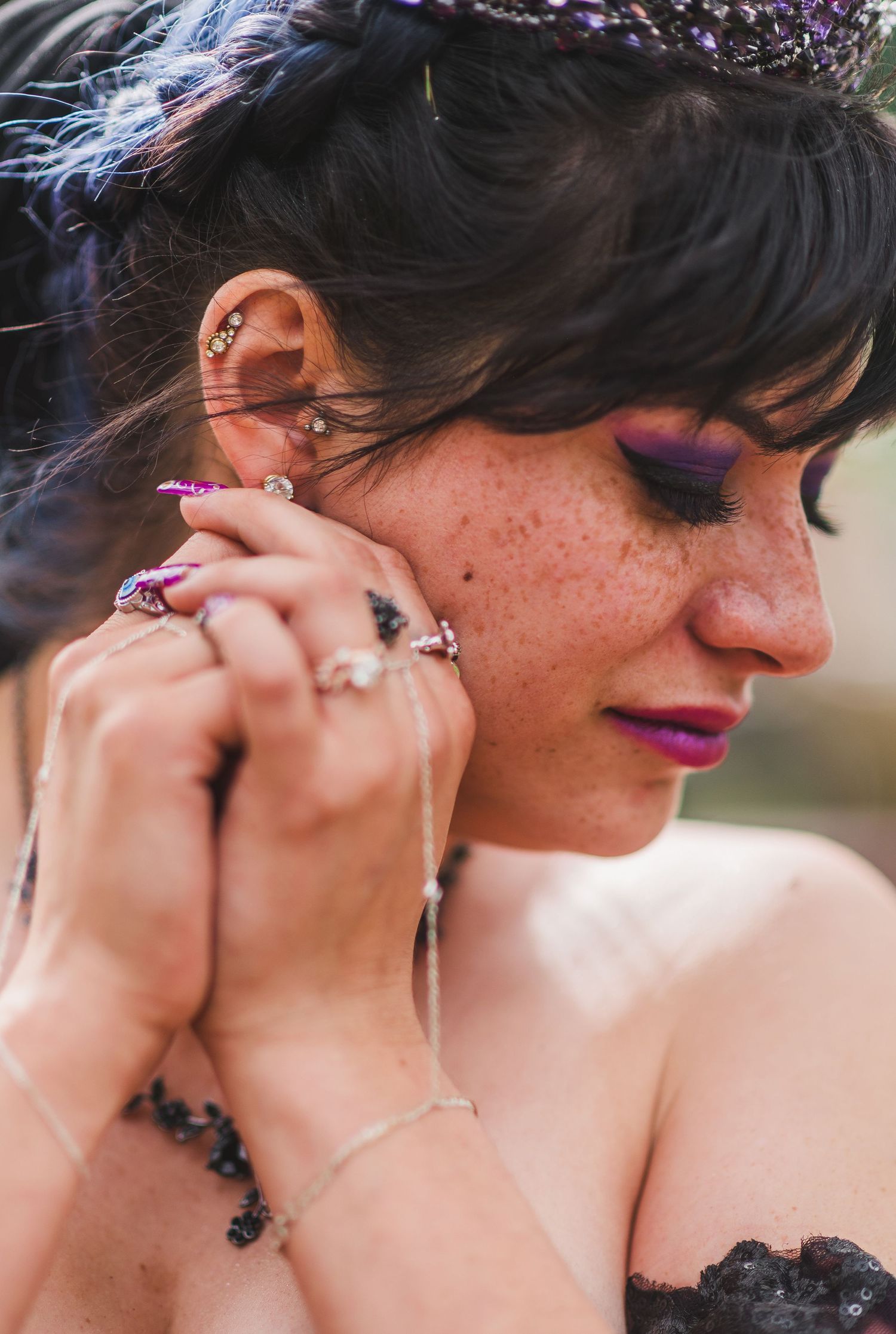 Woman with freckles, purple eye makeup, and multiple ear piercings adjusting earring, wearing lace top and hand chain bracelet.