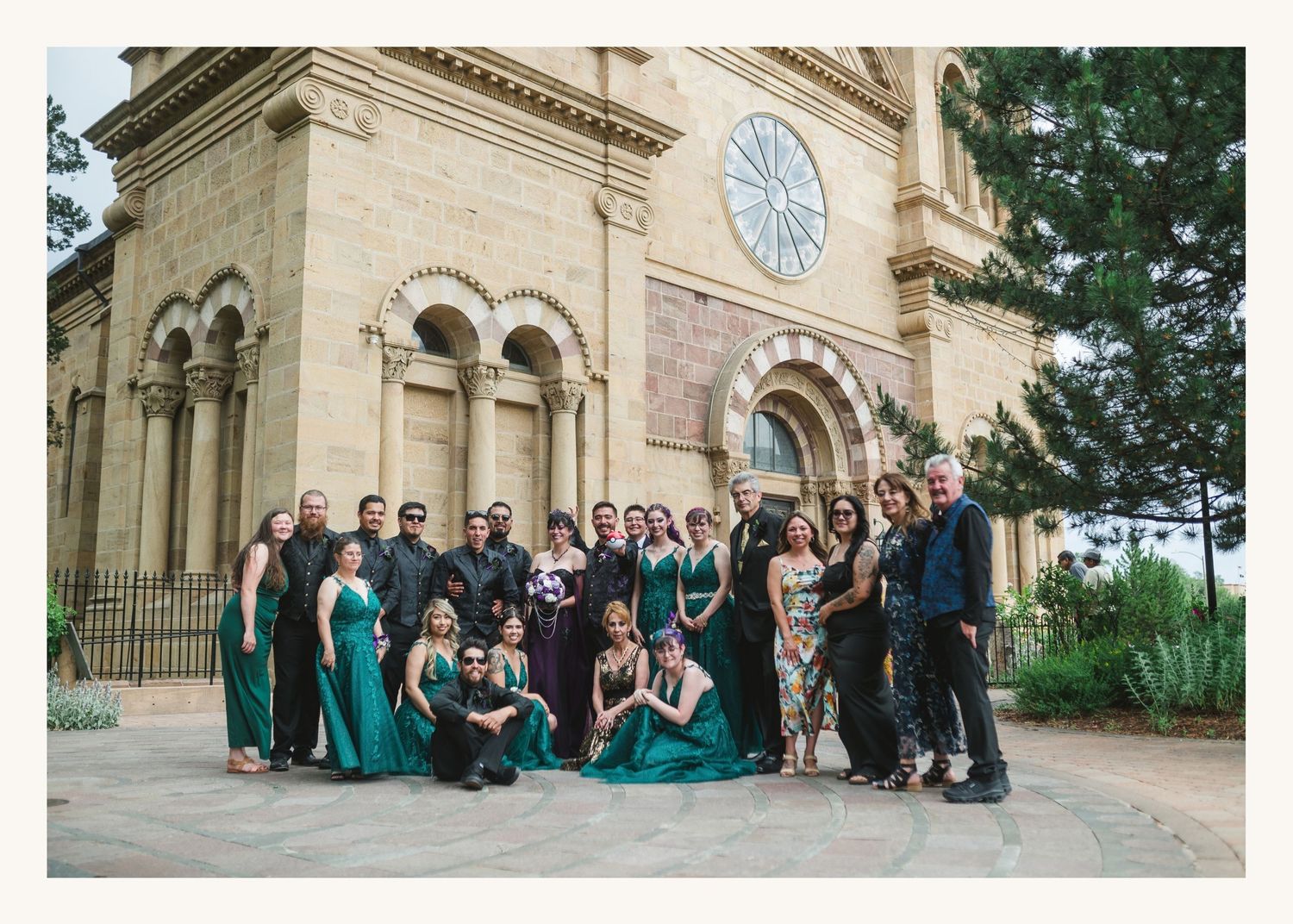 Large wedding party in emerald green dresses posed outside a historic stone cathedral in Santa Fe, NM.
