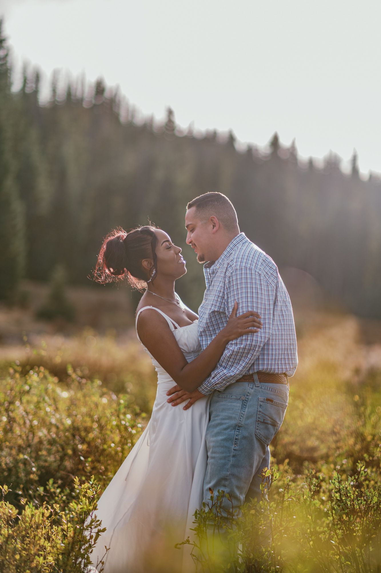 Couple embracing in a sunlit mountain meadow, woman in white dress and man in plaid shirt, surrounded by wildflowers.