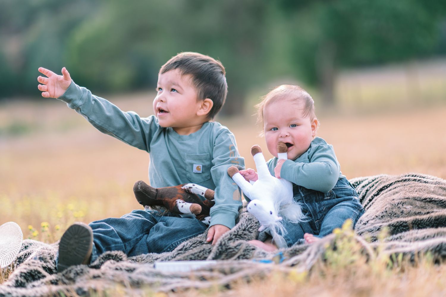 Two young toddlers sitting outdoors on a blanket in a field, one pointing excitedly while holding a stuffed unicorn toy.