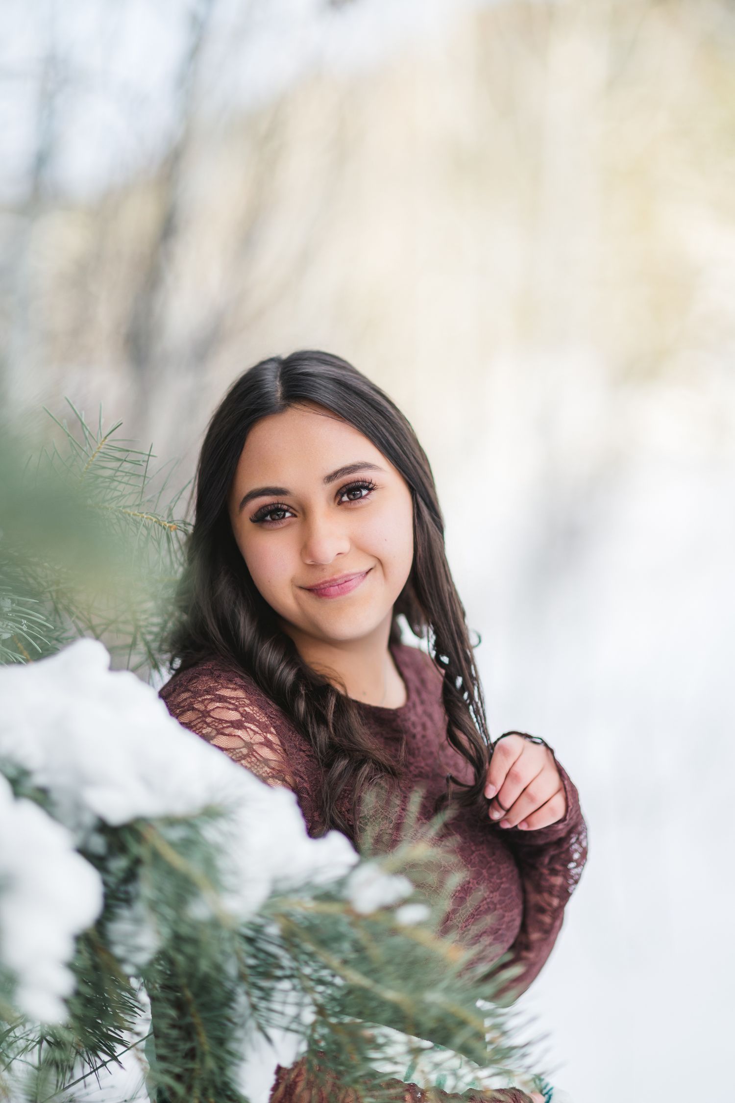 Portrait photo through snowy pine branches showing someone in a burgundy sweater smiling on a winter day.