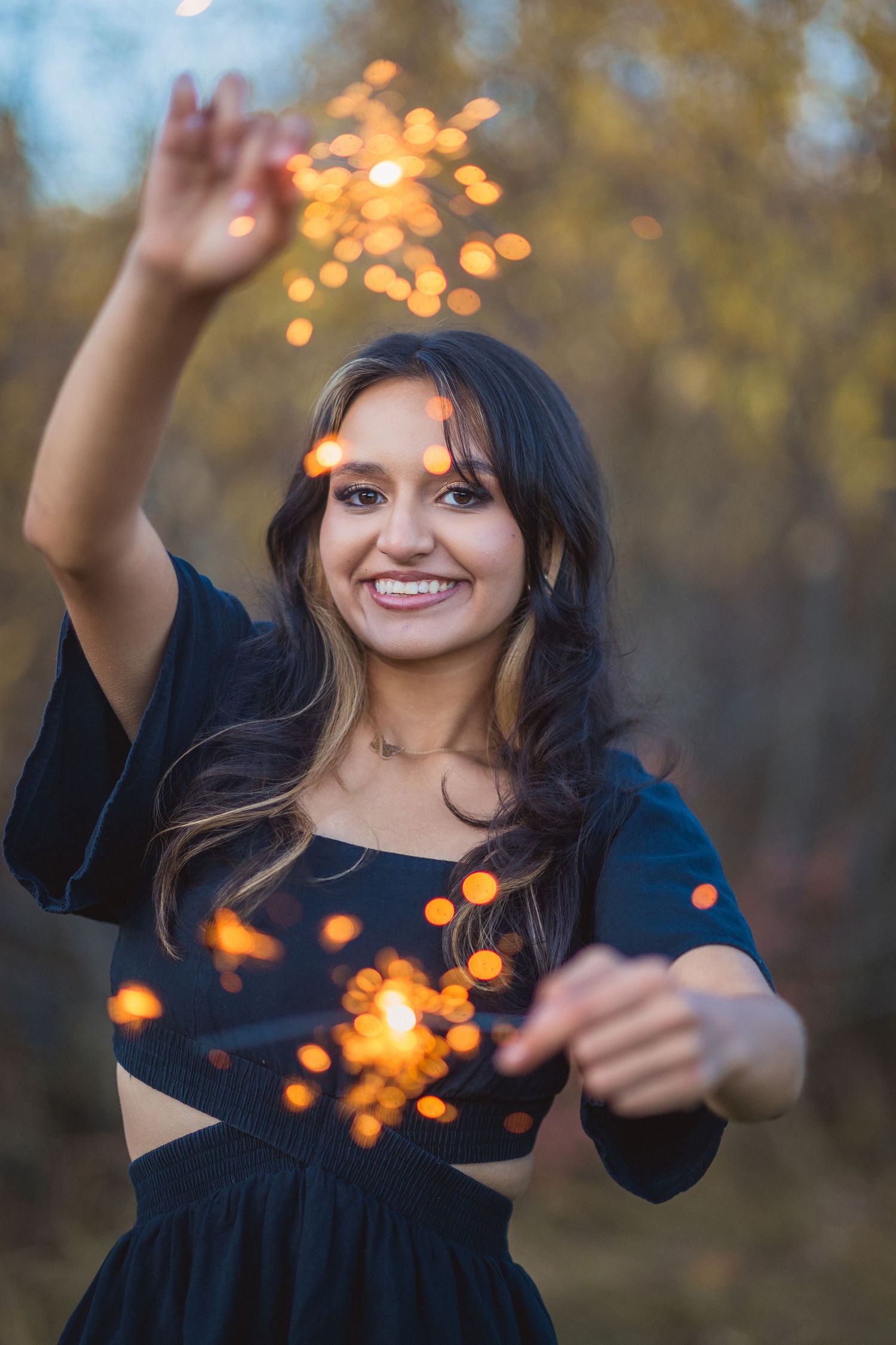 A joyful figure in a dark dress dances with sparklers against an autumn forest backdrop at dusk.