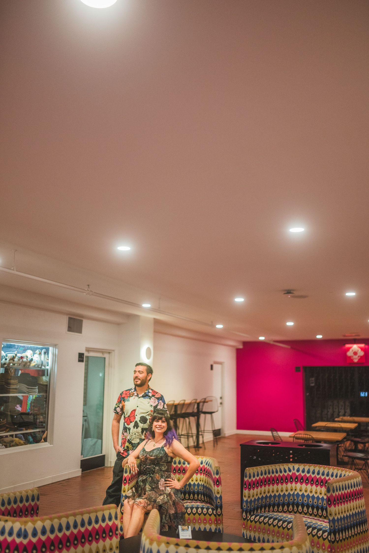 A couple poses in a vibrant lounge with colorful patterned sofas, recessed lighting, and a bright pink accent wall.