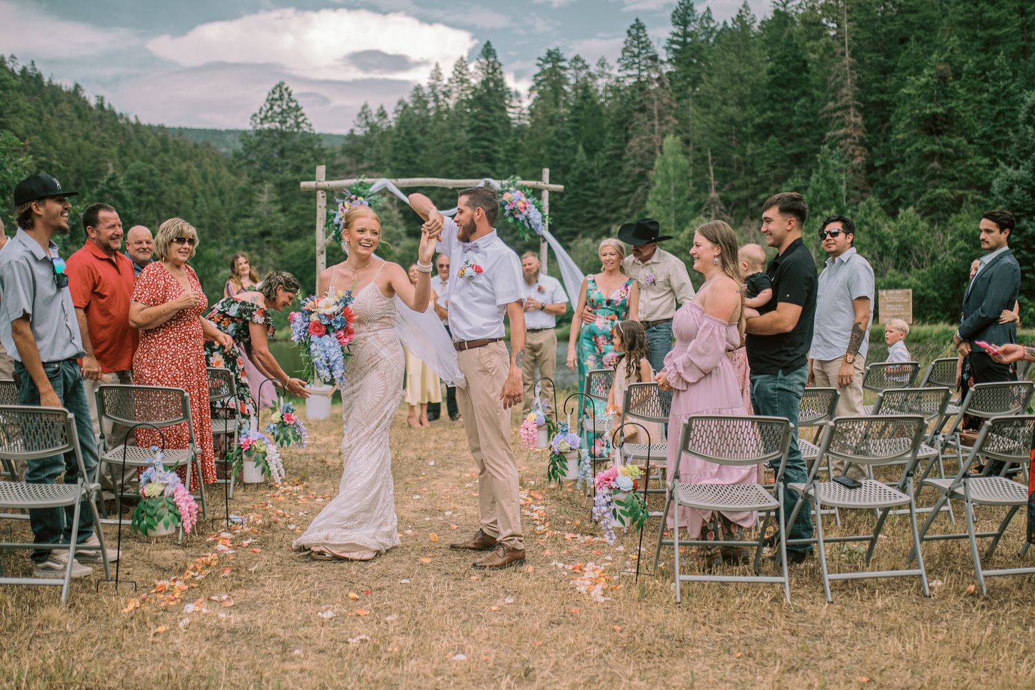 Outdoor wedding ceremony in forest clearing, bride and groom walking down aisle, guests celebrating, rustic arch backdrop.