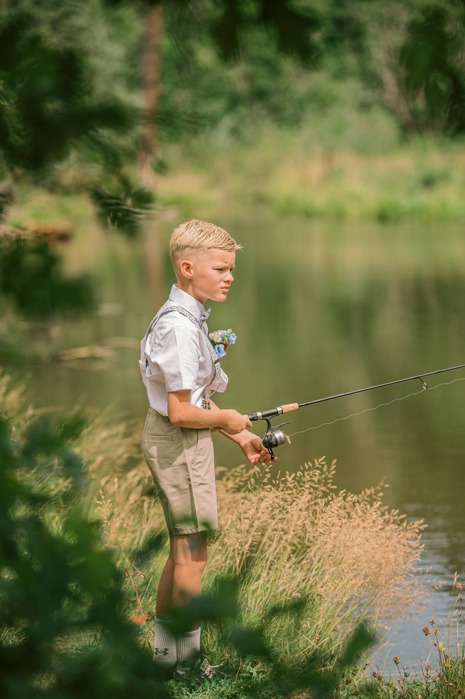Blonde boy fishing by a serene lake, holding a fishing rod among tall grass and lush green trees.