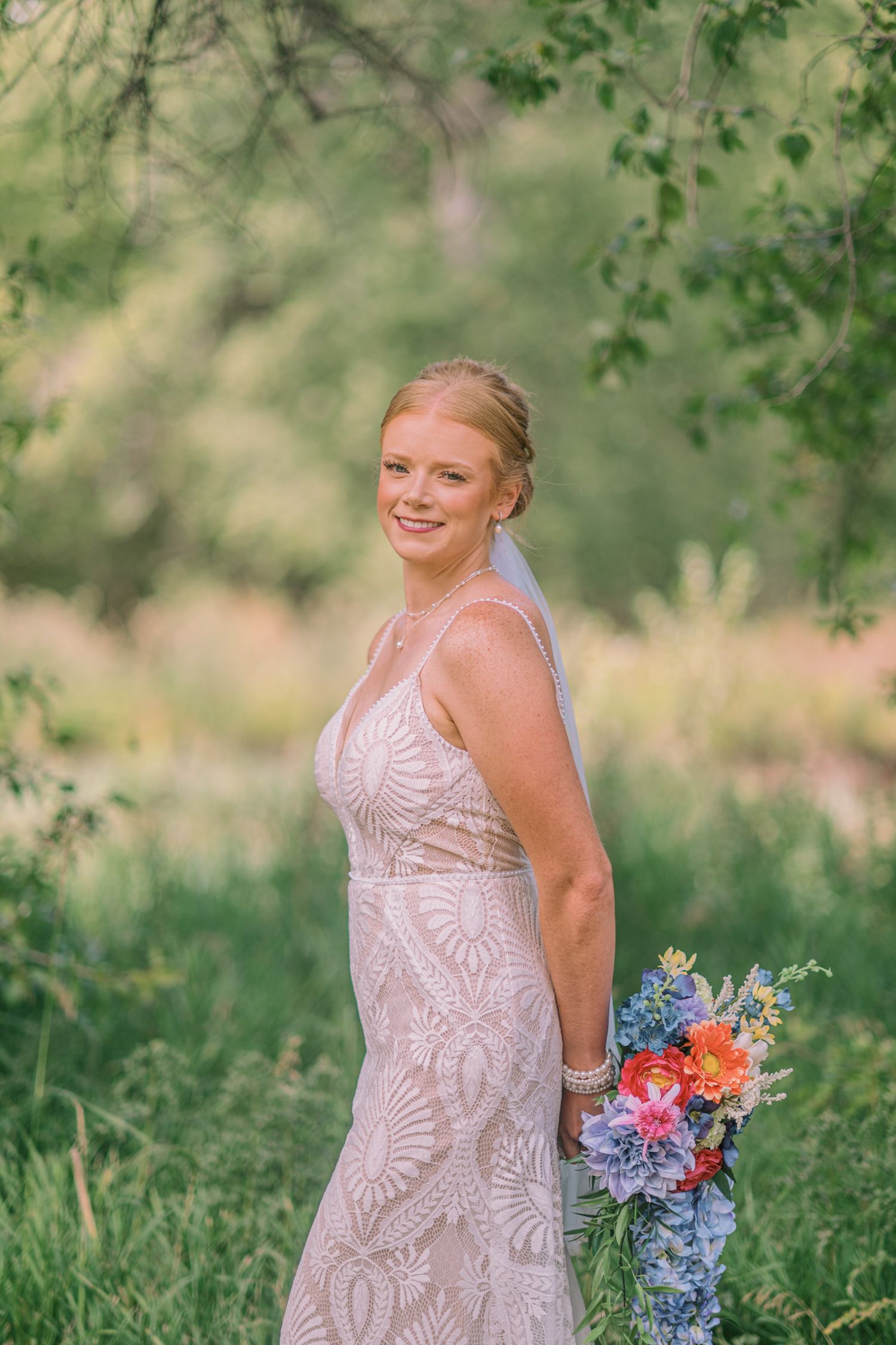 Smiling bride in lace gown holding colorful bouquet standing outdoors among lush green trees and meadow.