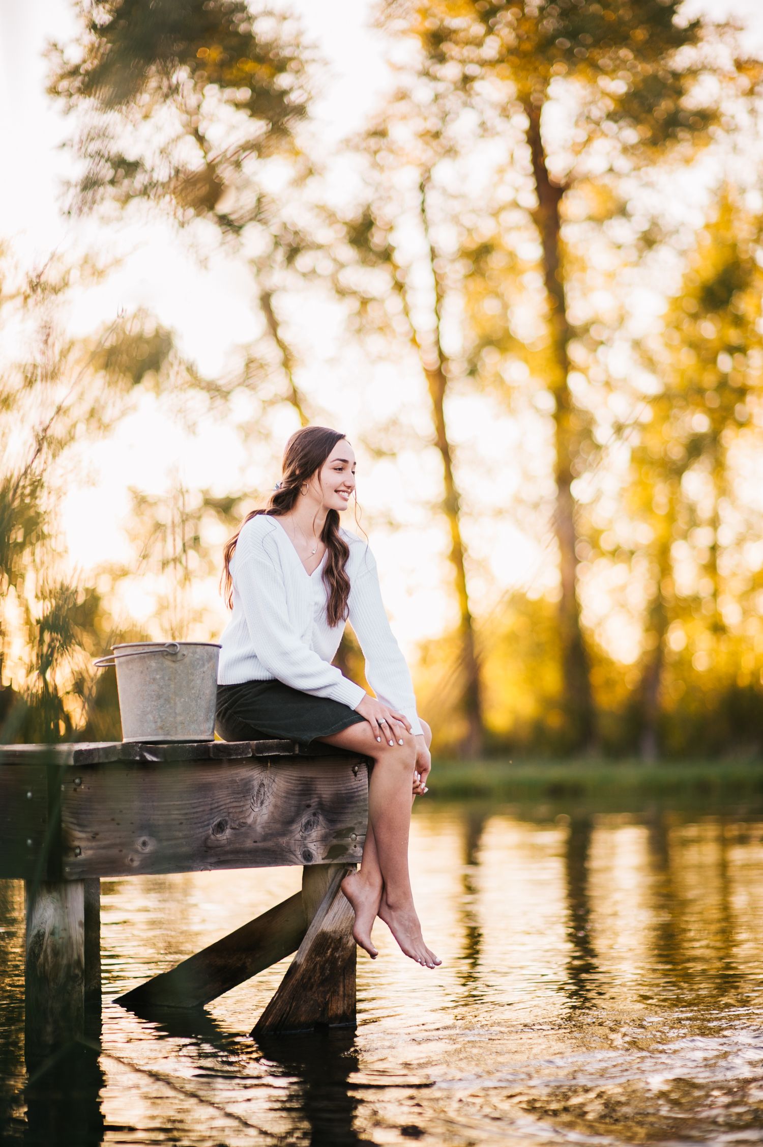 Person in white sweater seated on wooden pier over tranquil lake during sunset.