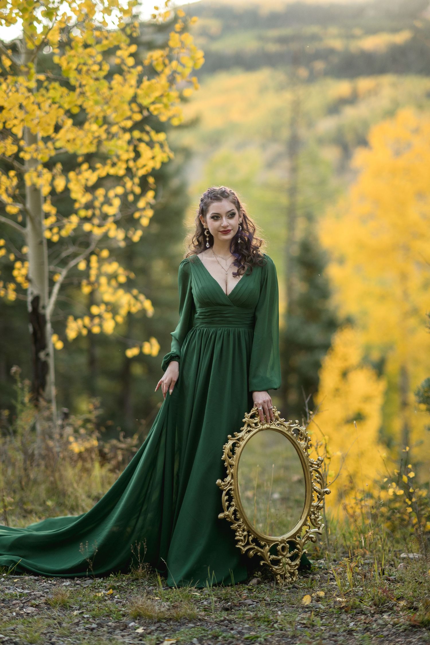 A woman in an emerald green dress poses gracefully among autumn trees with golden foliage in a dreamy outdoor setting.