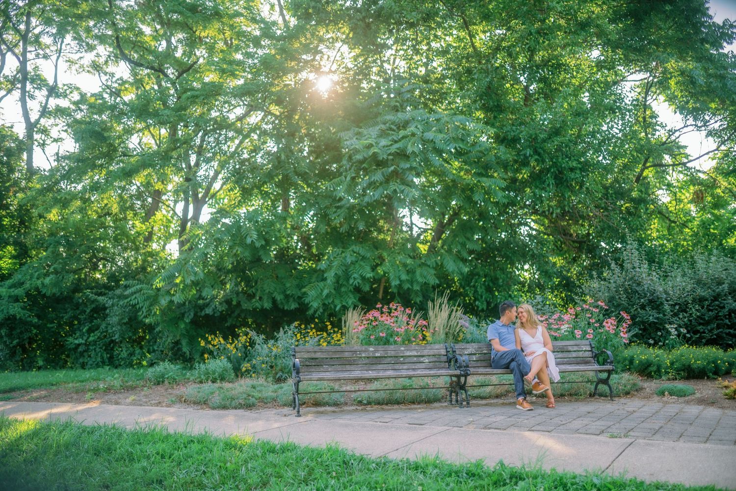 A person sits on a park bench surrounded by lush green trees and foliage with sunlight streaming through the branches.